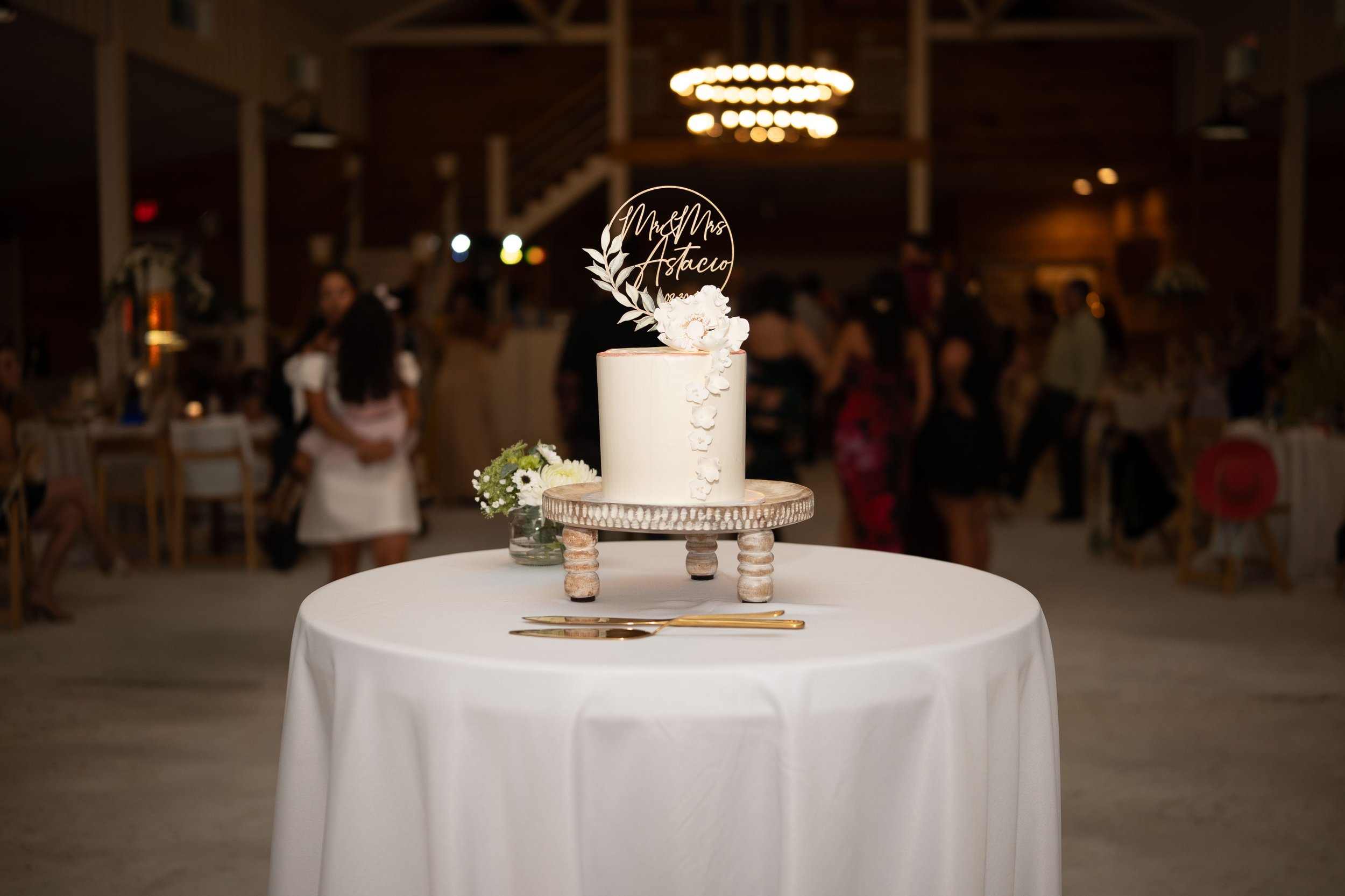 A white wedding cake on a round table with a white tablecloth at a wedding reception. The cake has a wooden topper with the words "Mr & Mrs. Astacio" and a floral decoration. In the background, people are mingling and dancing in a decorated rustic ve