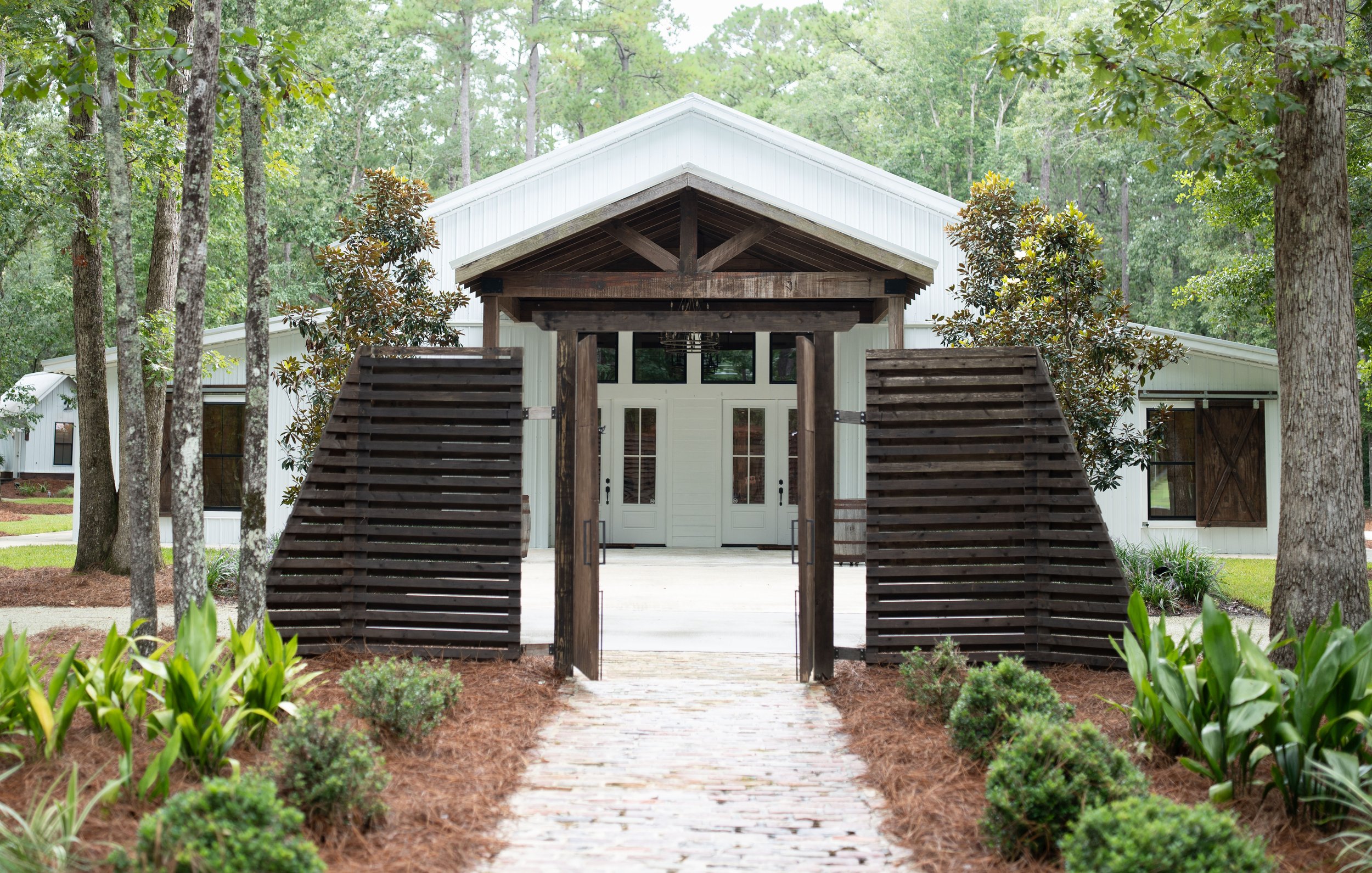 View of a white modern house with a wooden gate, surrounded by trees, plants, and a brick pathway leading to the front entrance.
