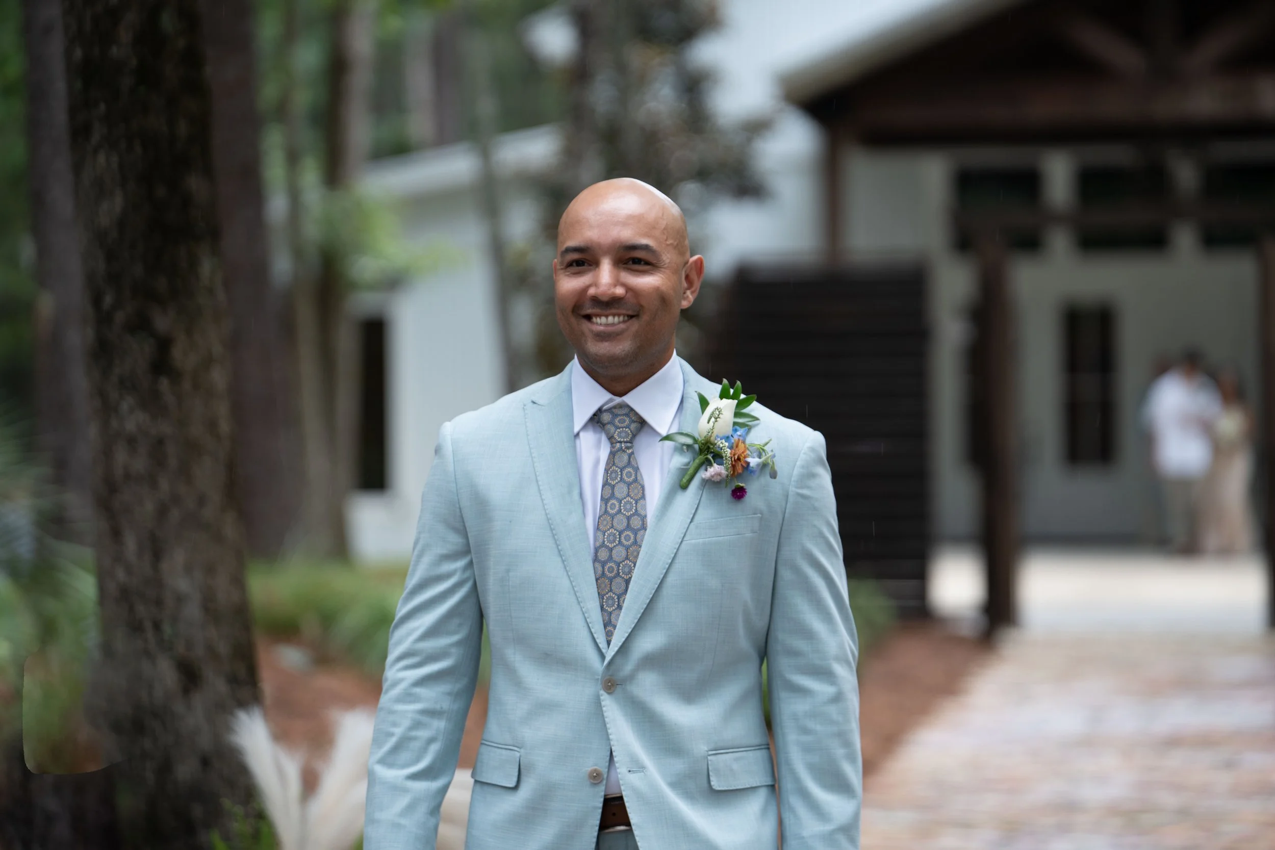 A man in a light blue suit with a floral boutonniere smiling outdoors, with trees and a building in the background.