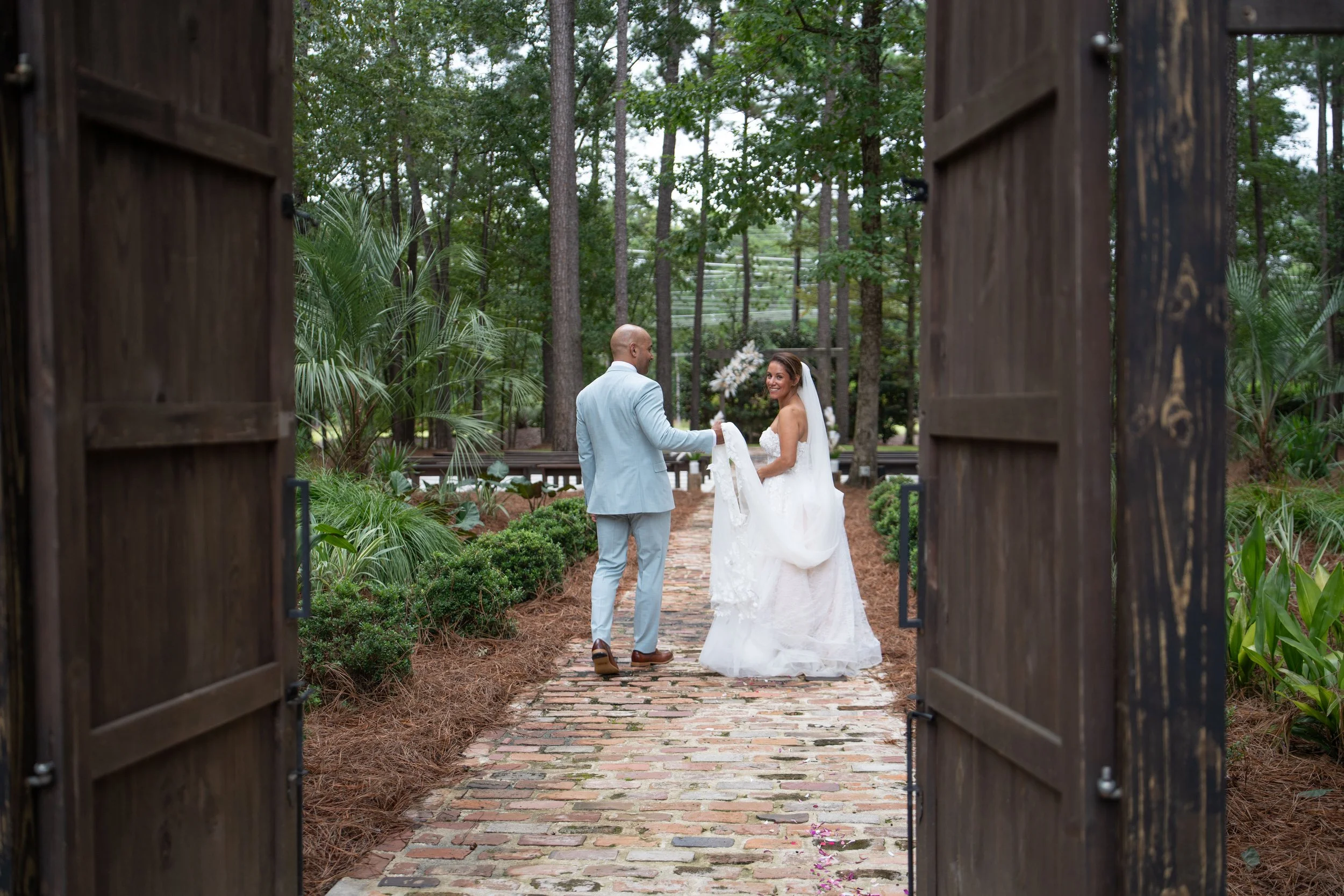 A bride and groom holding hands outdoors in a wooded setting during their wedding, viewed through an open wooden gate.
