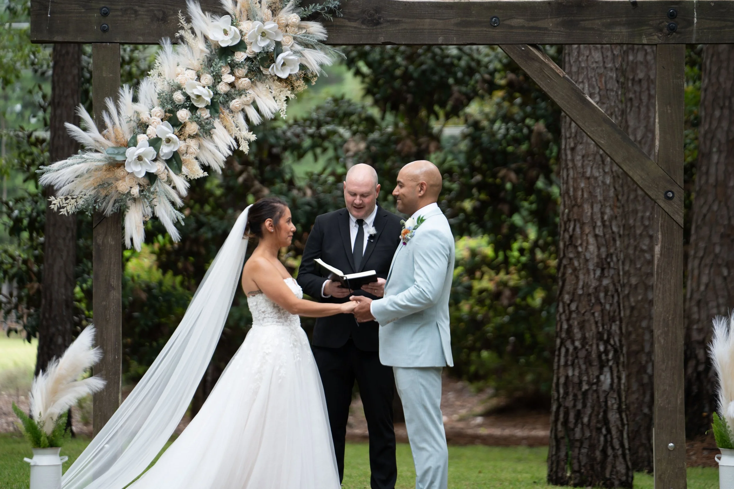 A wedding ceremony taking place outdoors under a decorated arch with the bride and groom holding hands, exchanging vows, with an officiant standing between them, surrounded by trees.