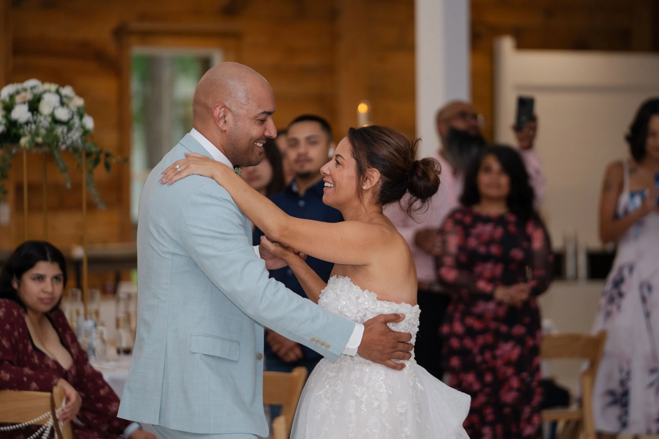 A couple dancing at their wedding reception, the bride and groom smiling at each other, surrounded by guests in a wooden hall.