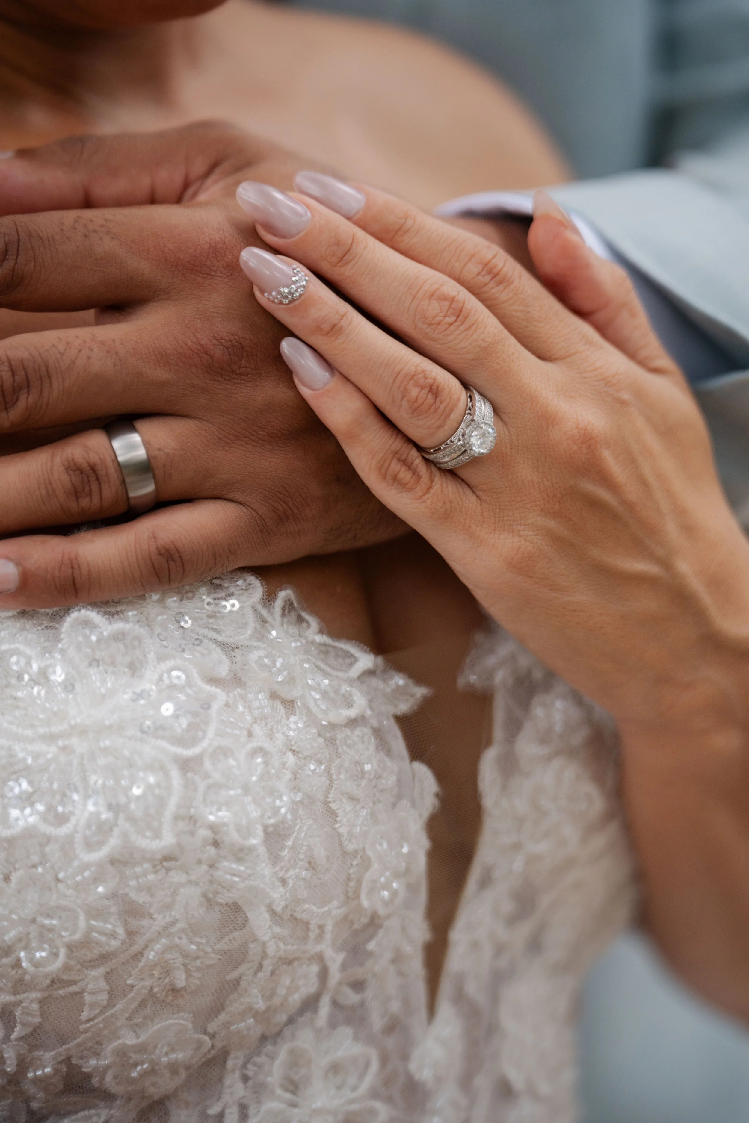 Close-up of a bride's hand on a groom's chest, showing wedding rings and intricate lace wedding dress.