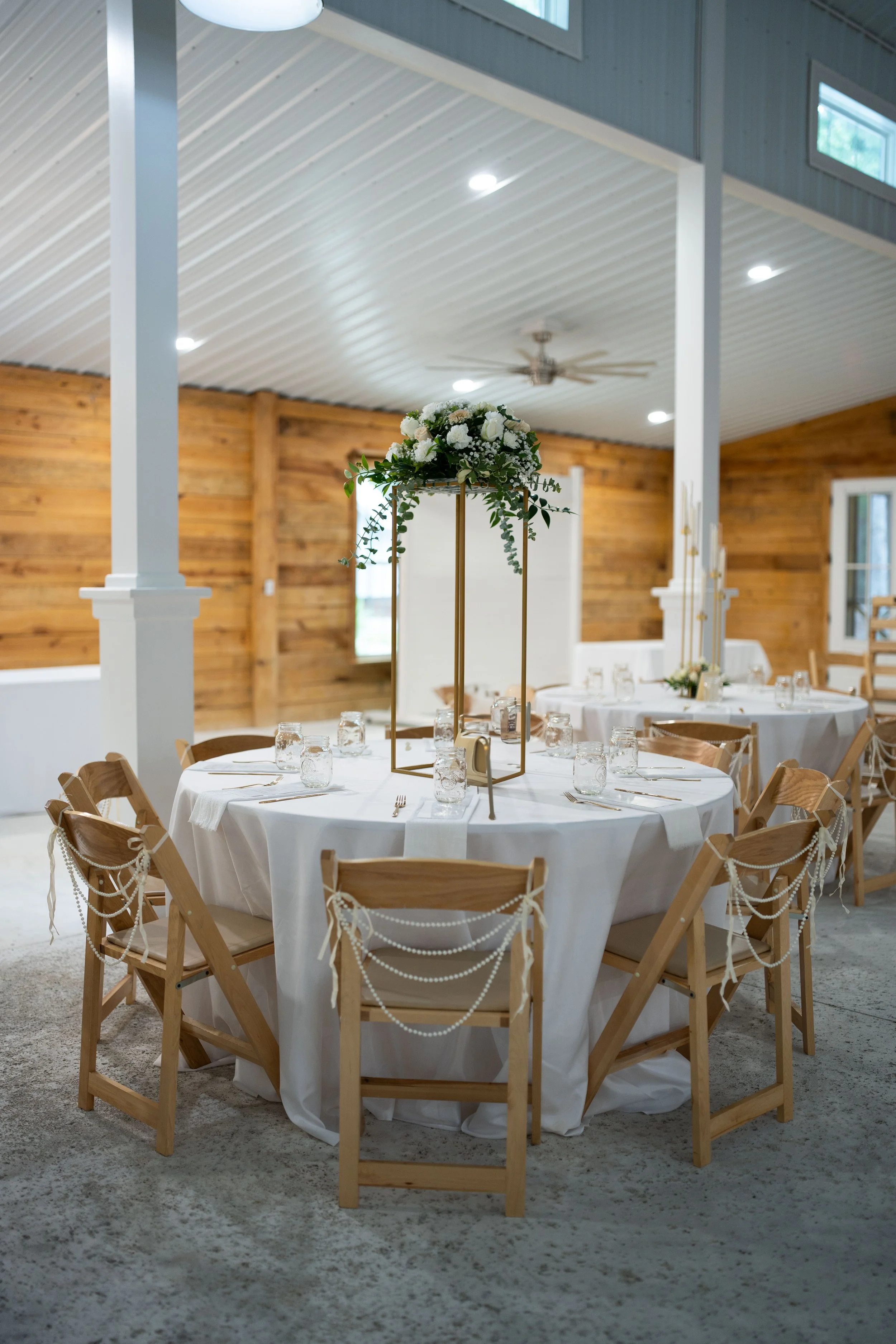 Round table set for a formal event with white tablecloth, glass jars for drinks, silverware, and a floral centerpiece with white roses and greenery, in a rustic venue with wood-paneled walls and a ceiling fan.