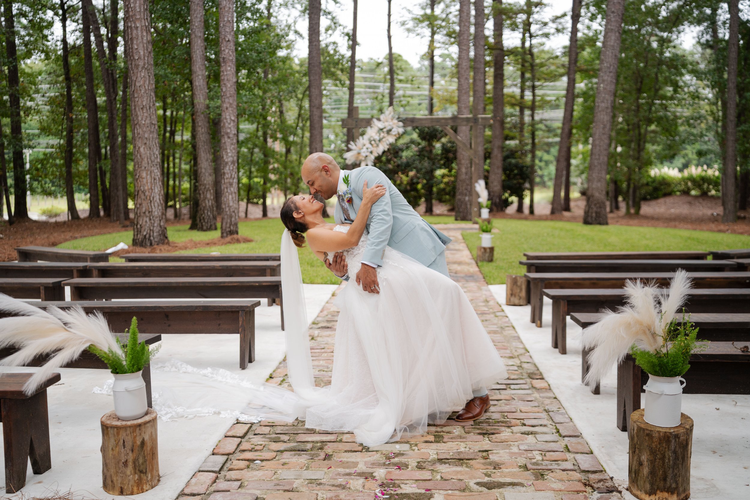 A bride and groom share a joyful moment during their outdoor wedding ceremony in a forested area, with benches and floral decorations.