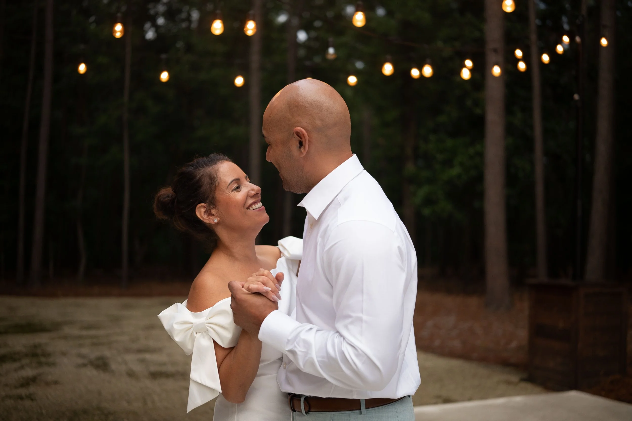 A couple dancing and smiling at an outdoor wedding reception at dusk, with string lights hanging in a wooded area in the background.
