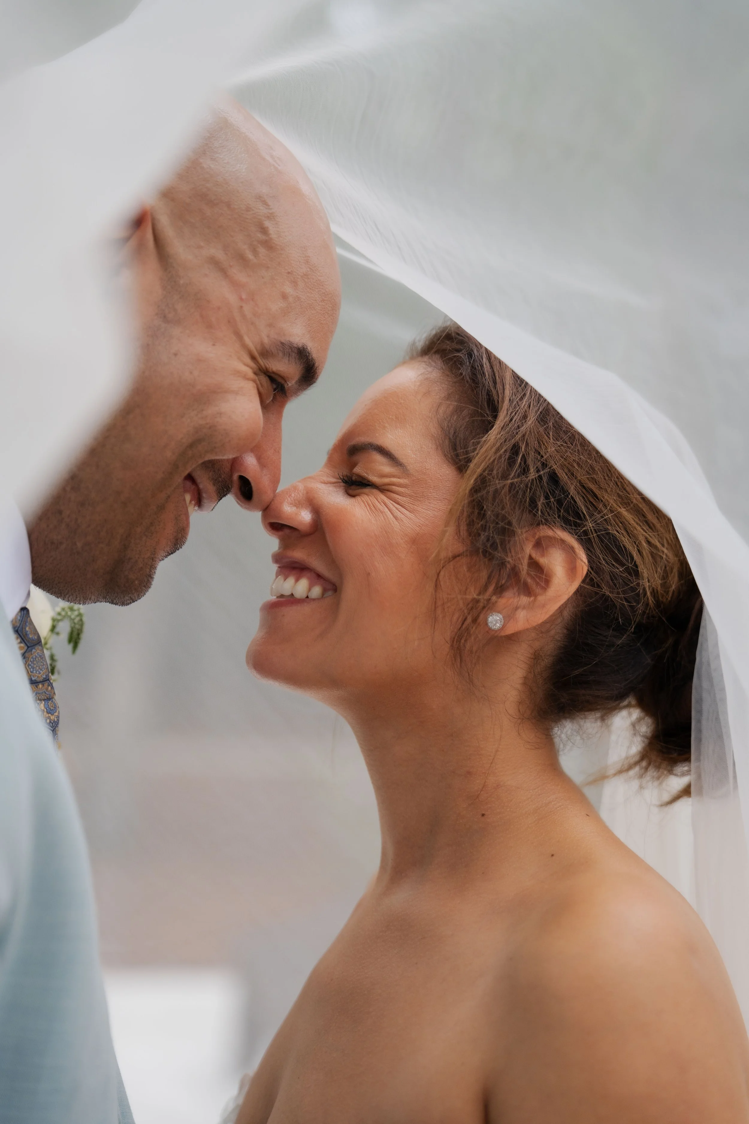 A bride and groom smiling and touching noses under a white veil during their wedding.