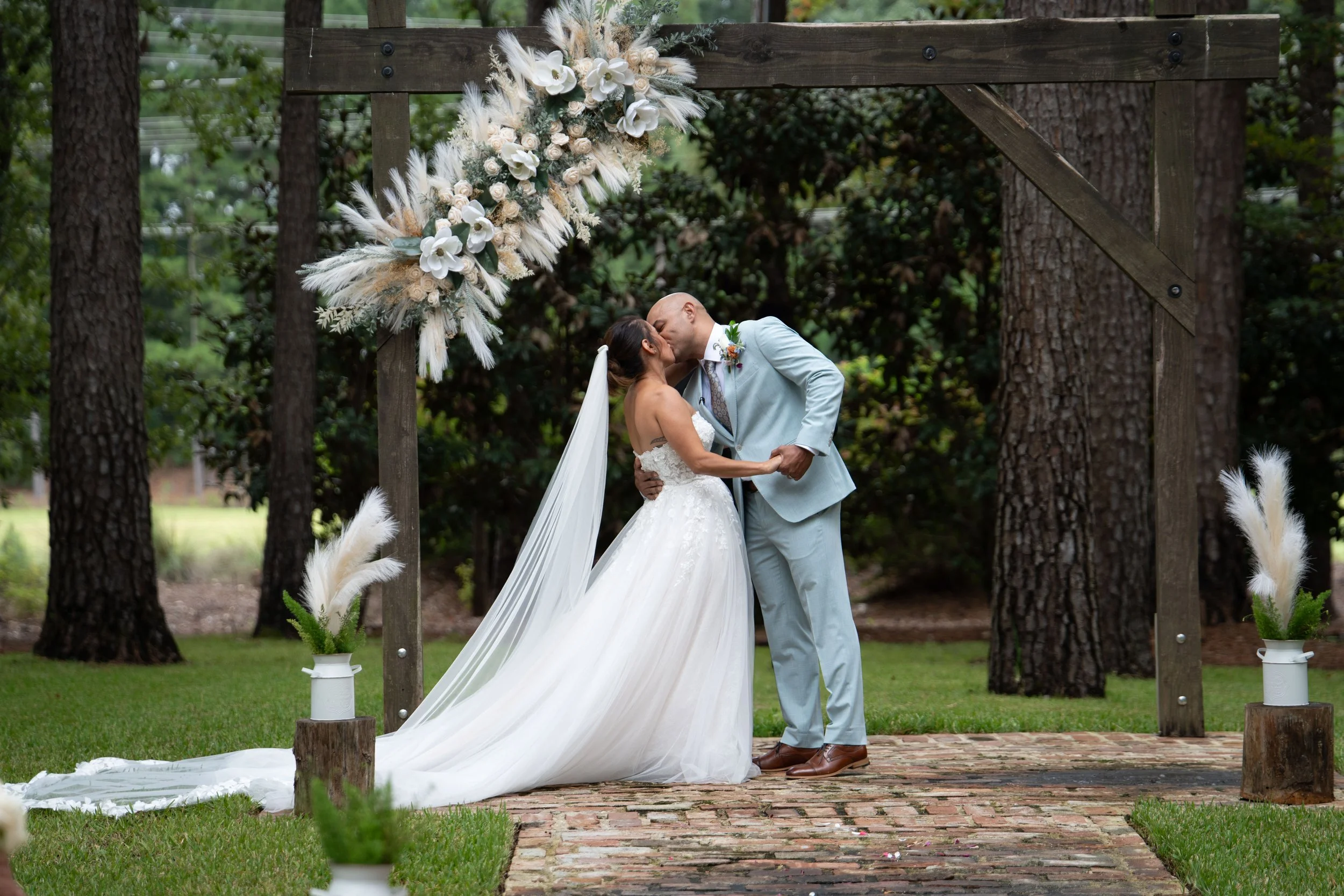 A bride and groom sharing a kiss during their outdoor wedding ceremony under a wooden arch decorated with white and cream flowers and pampas grass, with potted plants on either side, in a wooded park.
