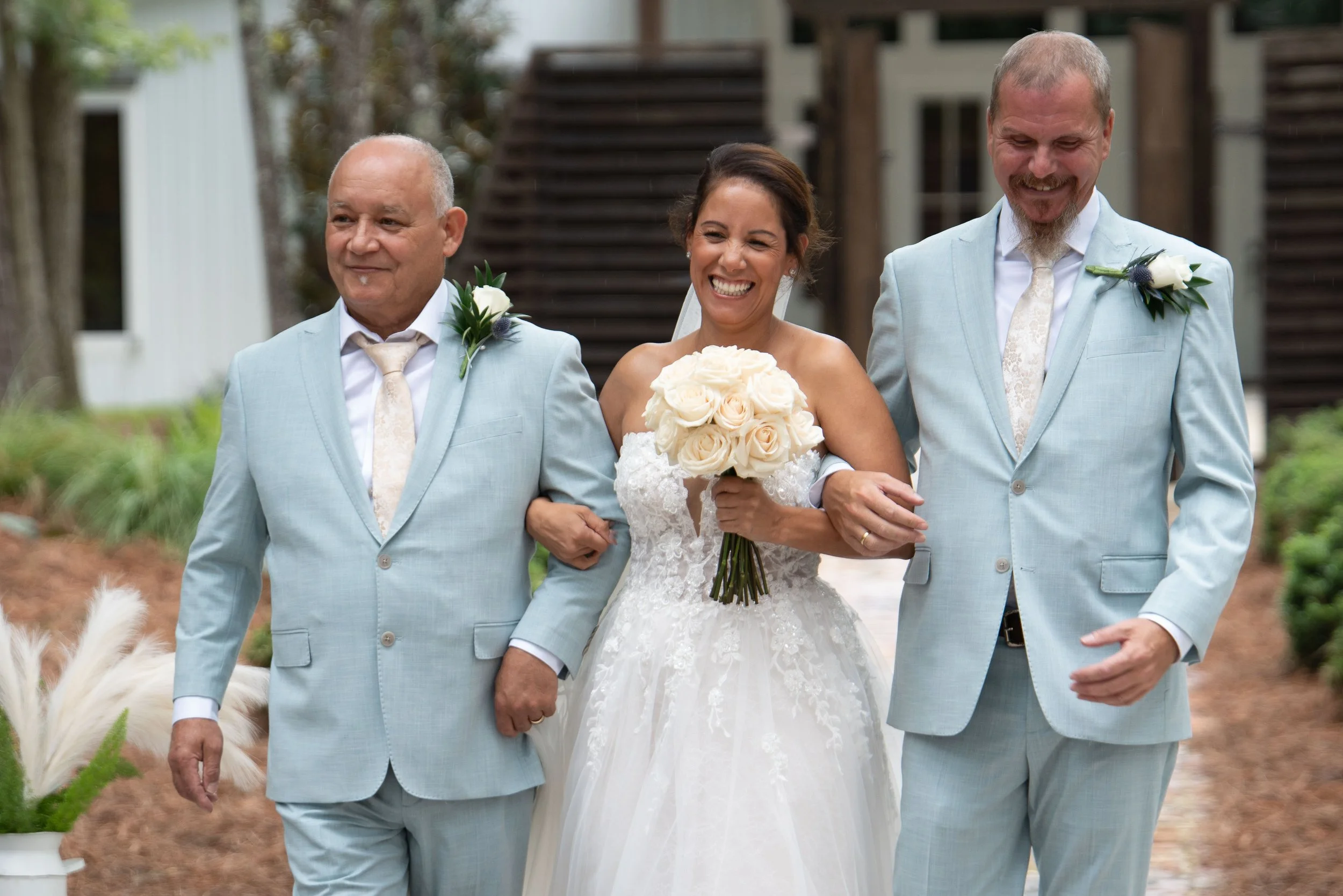 A bride in a white wedding gown holding a bouquet of white roses, walking arm-in-arm with two men in light blue suits, outdoor wedding setting with greenery and a building in the background.