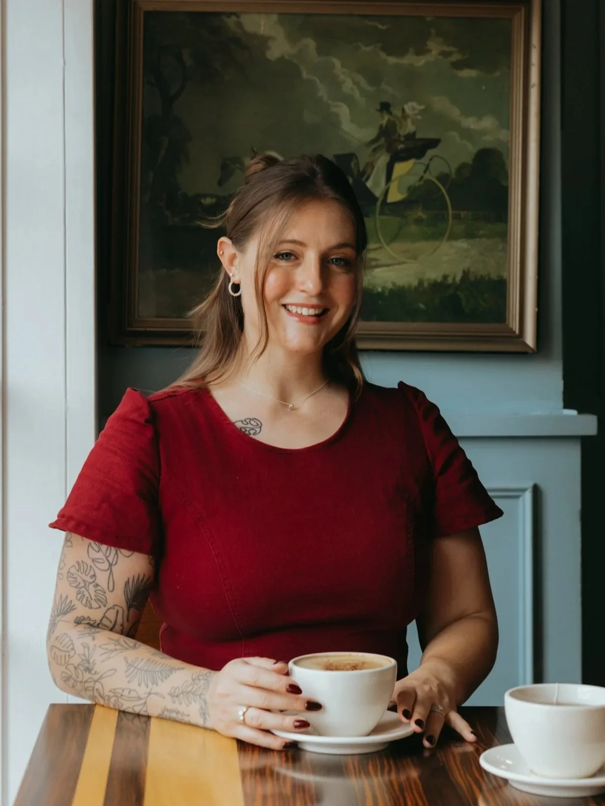 A woman with tattoos and earrings, wearing a red top, sits at a table in a cafe, smiling and holding a cup of coffee or tea. There is another cup on the table and a painting of a person riding a horse-drawn carriage hanging on the wall behind her.