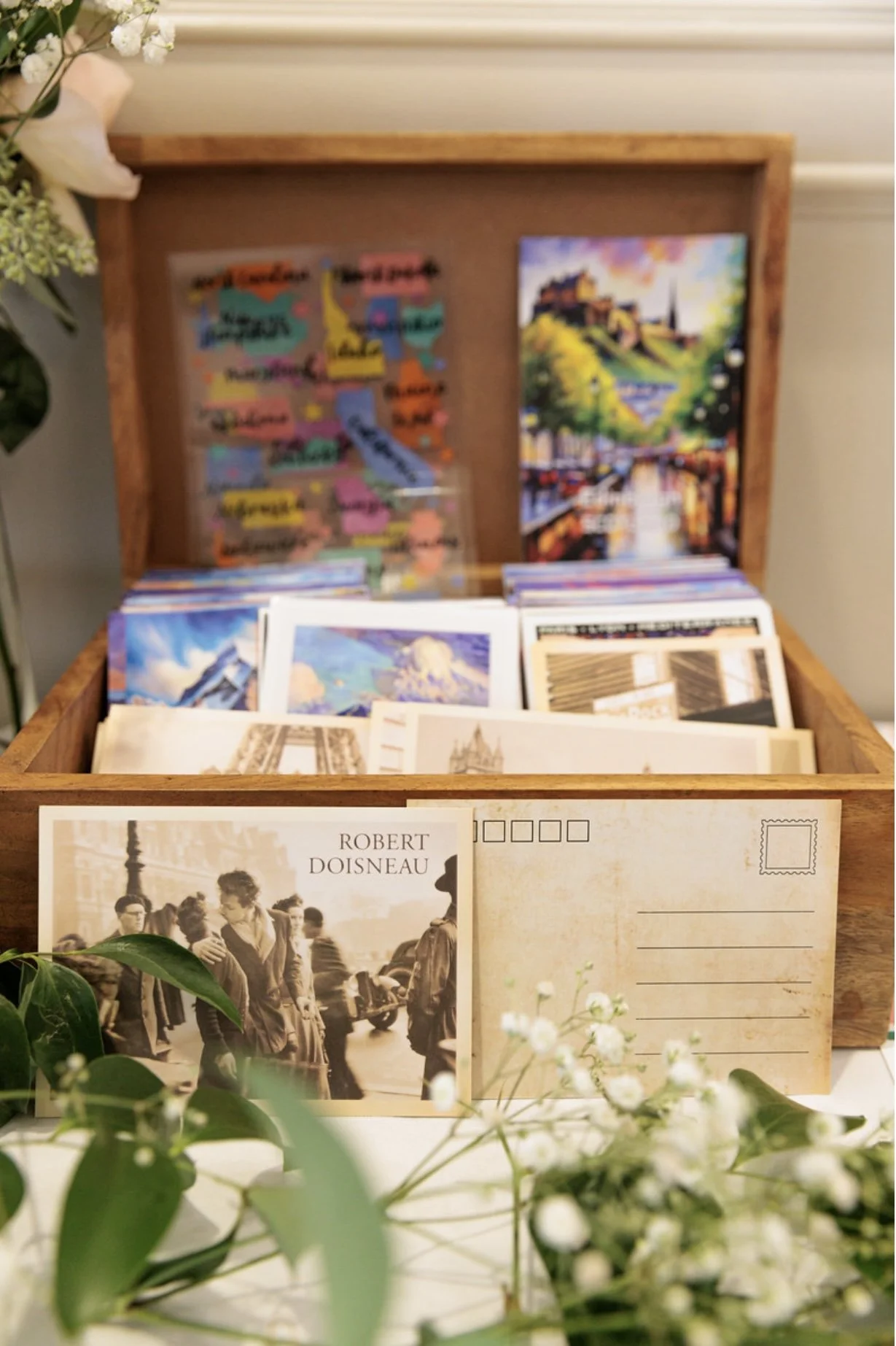 A wooden display box filled with colorful postcards and art prints, with a postcard featuring Robert Doisneau's photograph of people on a Paris street in the foreground. There are flowers and greenery in front of the box.