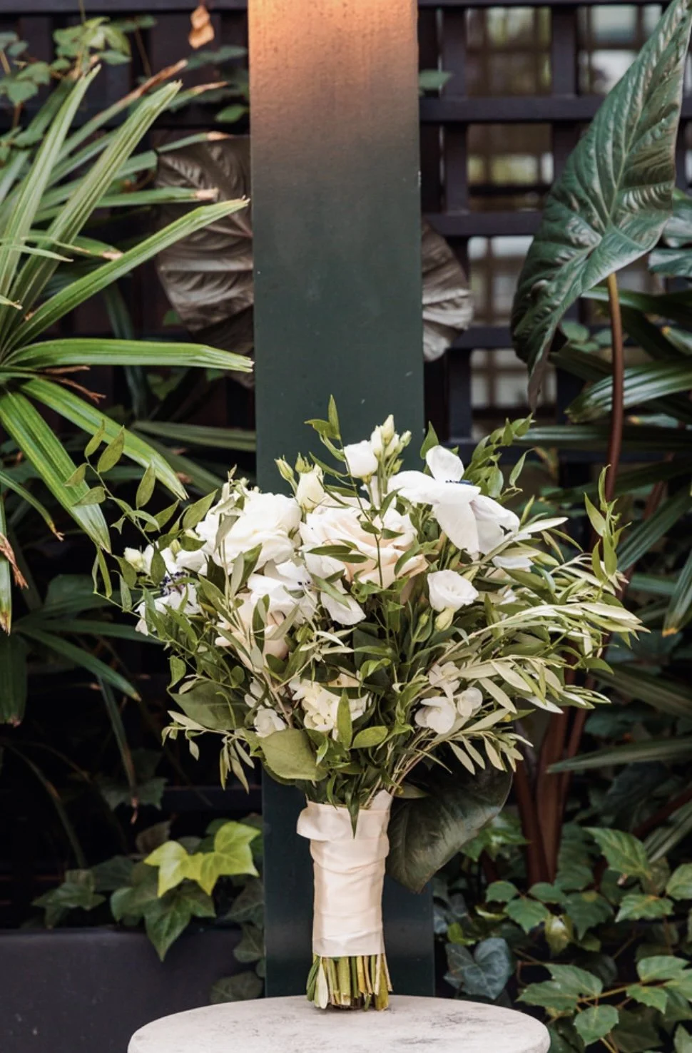 A bouquet of white flowers and green foliage wrapped in cream-colored cloth, placed on a light-colored surface, with various green plants in the background.