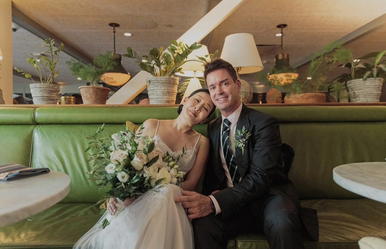 A bride and groom sitting on a green booth in a restaurant, smiling and hugging. The bride is holding a bouquet of white and purple flowers, wearing a lace wedding dress. The groom is dressed in a dark suit with a striped tie.