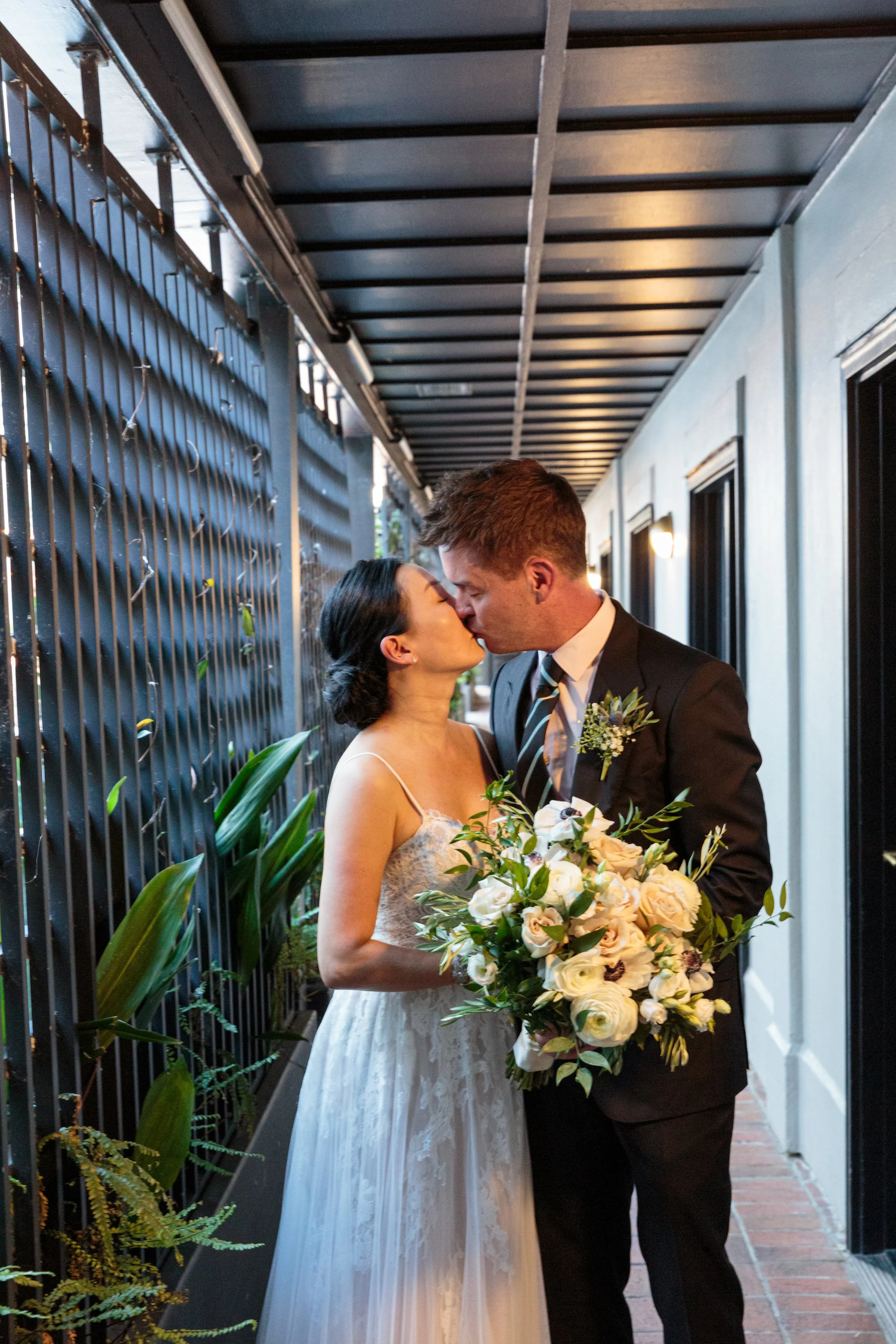 A bride and groom share a kiss on their wedding day, with the bride holding a large bouquet of white and cream flowers, standing under a covered outdoor corridor with plants along the side.