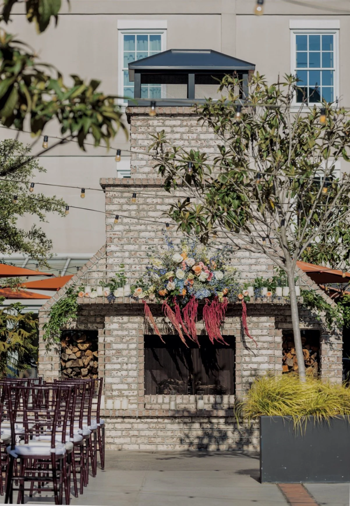 Outdoor scene featuring a brick fireplace with a colorful floral arrangement on top, surrounded by trees, string lights, and chairs with white cushions.