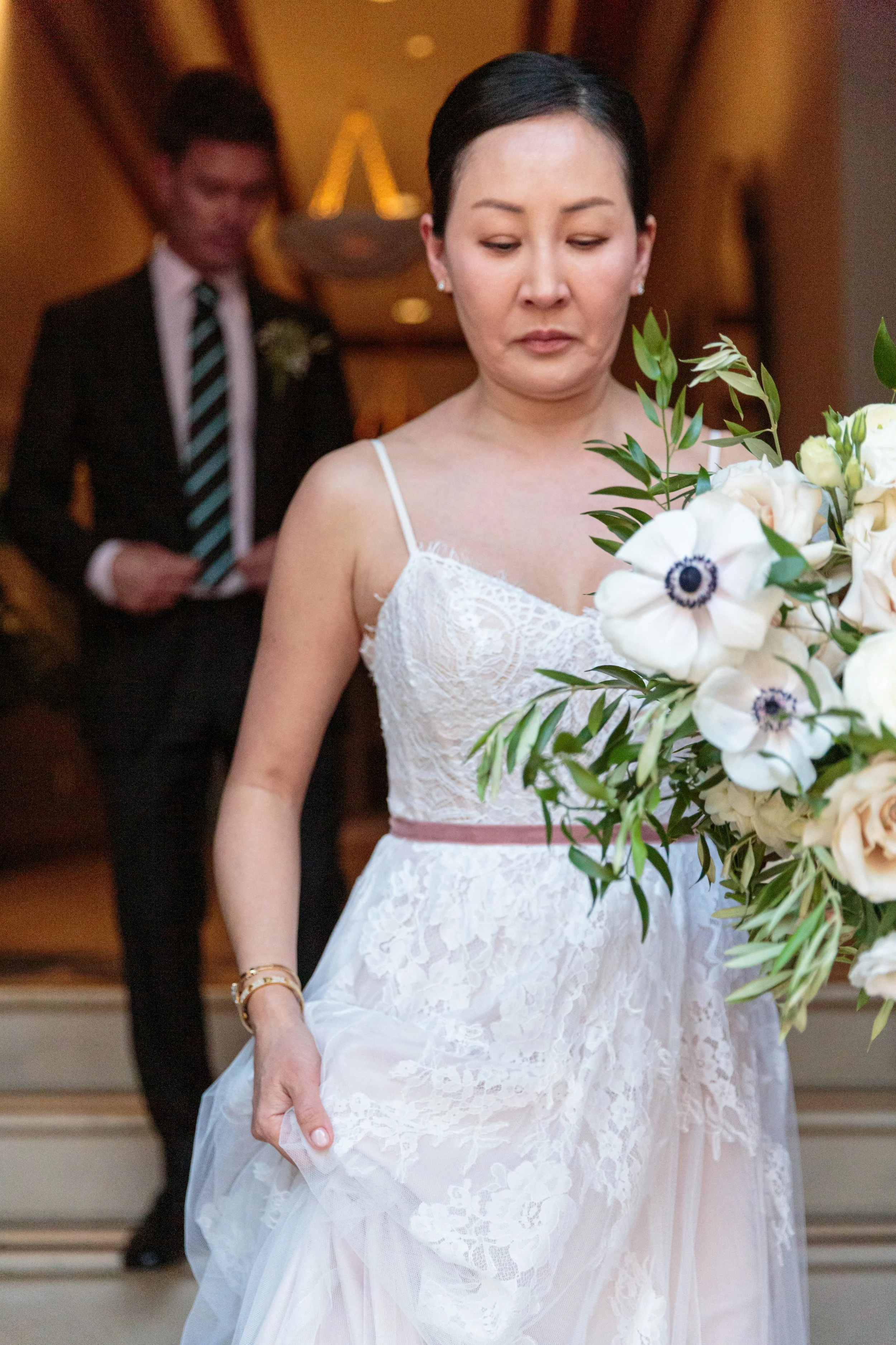 A woman in a wedding dress holding a bouquet of white and blush roses, with a man in a suit in the background.