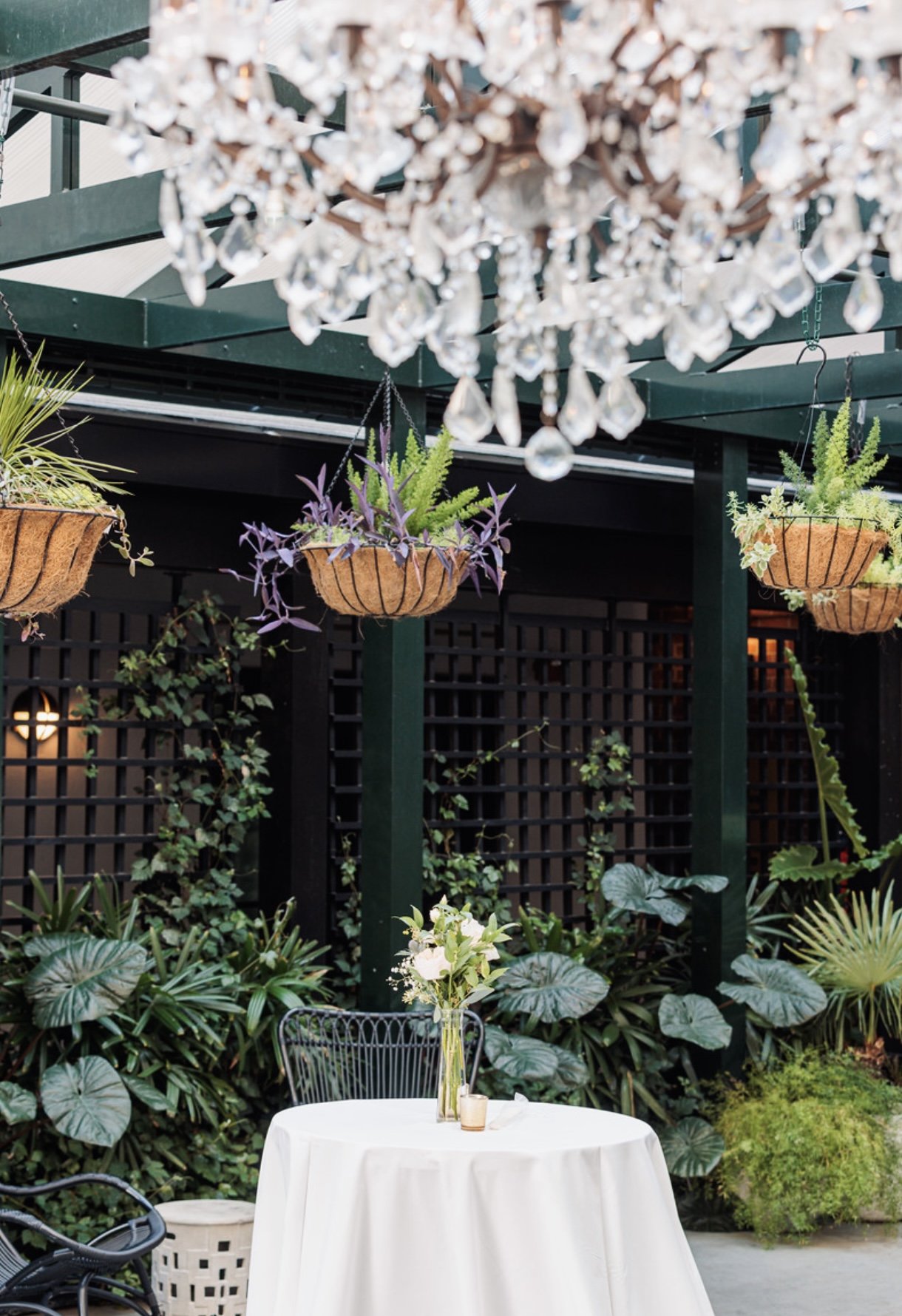 Outdoor patio with a round table covered with a white tablecloth, a vase with white flowers, and a small candle. Hanging planters with purple and green plants, a crystal chandelier hanging above, and lush green foliage in the background.