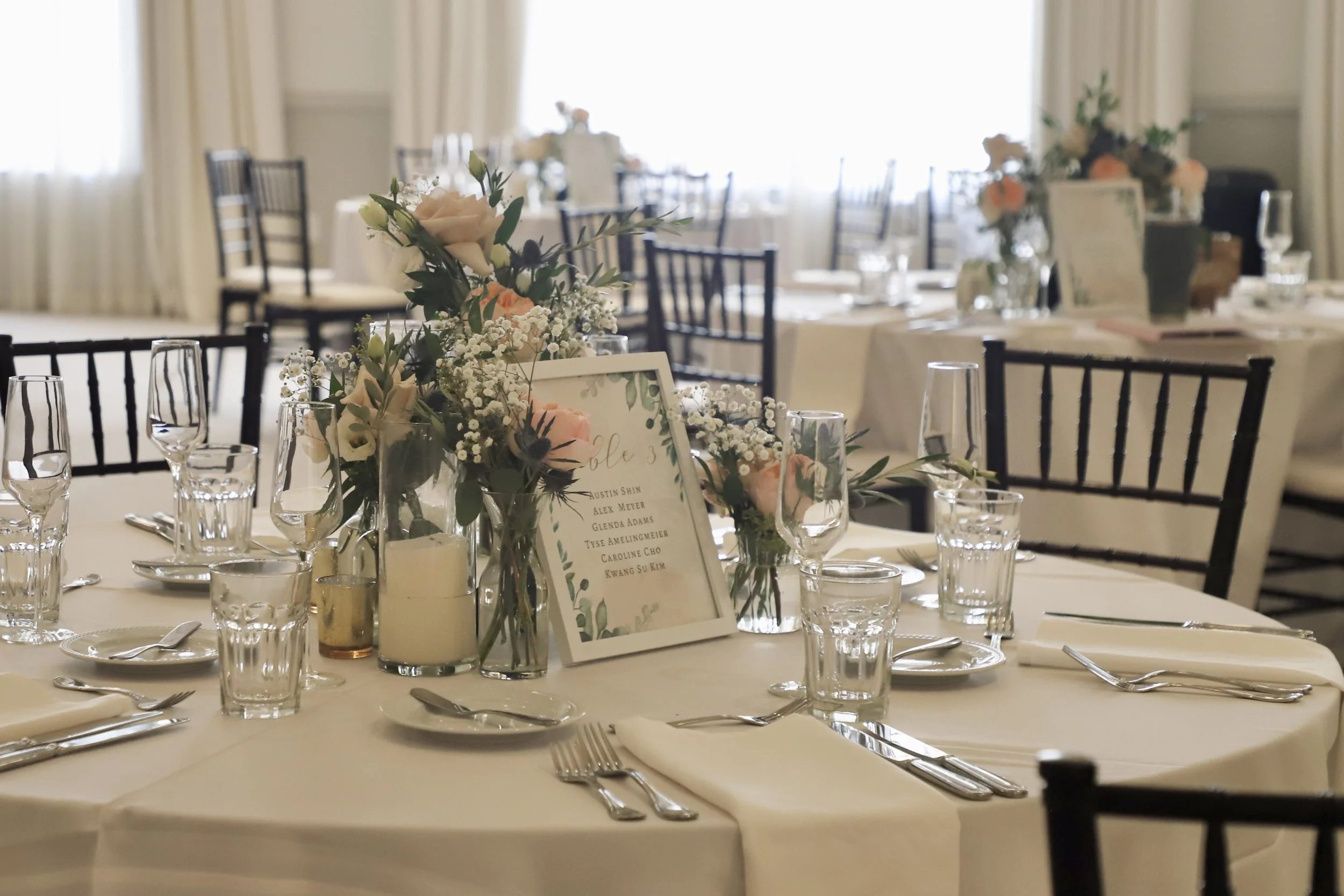 Elegant round banquet table set for a formal event with white tablecloth, floral centerpiece, glassware, silverware, and a framed table number or menu card in a well-lit room with large windows and black chairs.