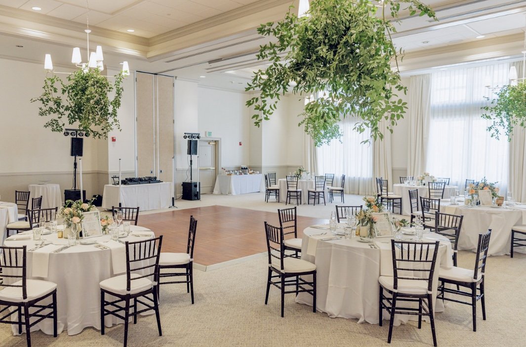 Event space with round tables covered in white tablecloths and black chairs, centerpieces with flowers, a small dance floor, and hanging green plants, with windows and curtains in the background.
