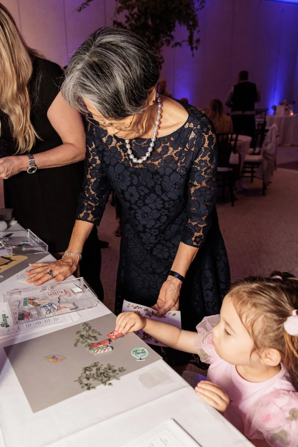 An elderly woman with short gray hair, glasses, wearing a navy blue lace dress and pearls, is leaning over a table, showing a Christmas-themed card to a young girl with light brown hair, pink dress, and a pink bow in her hair, at a festive event.