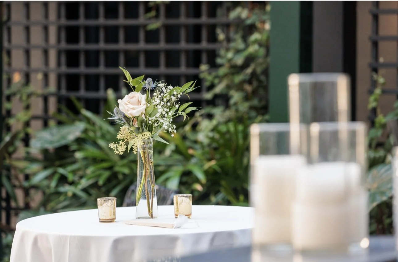 A round table with a white tablecloth, decorated with a floral centerpiece in a clear glass vase and small candles in glass holders, outdoors with lush greenery and a wooden lattice background.