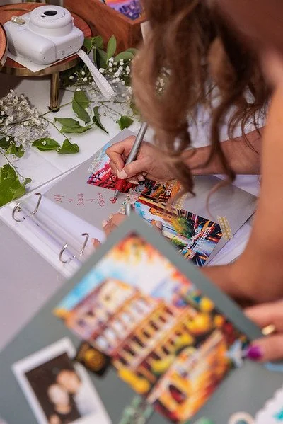 A woman with curly hair signs a book with a silver marker in a scrapbook-style photo album. There are decorative flowers and a camera on the table, along with a colorful postcard or magazine page. A small polaroid-style photo is visible at the bottom