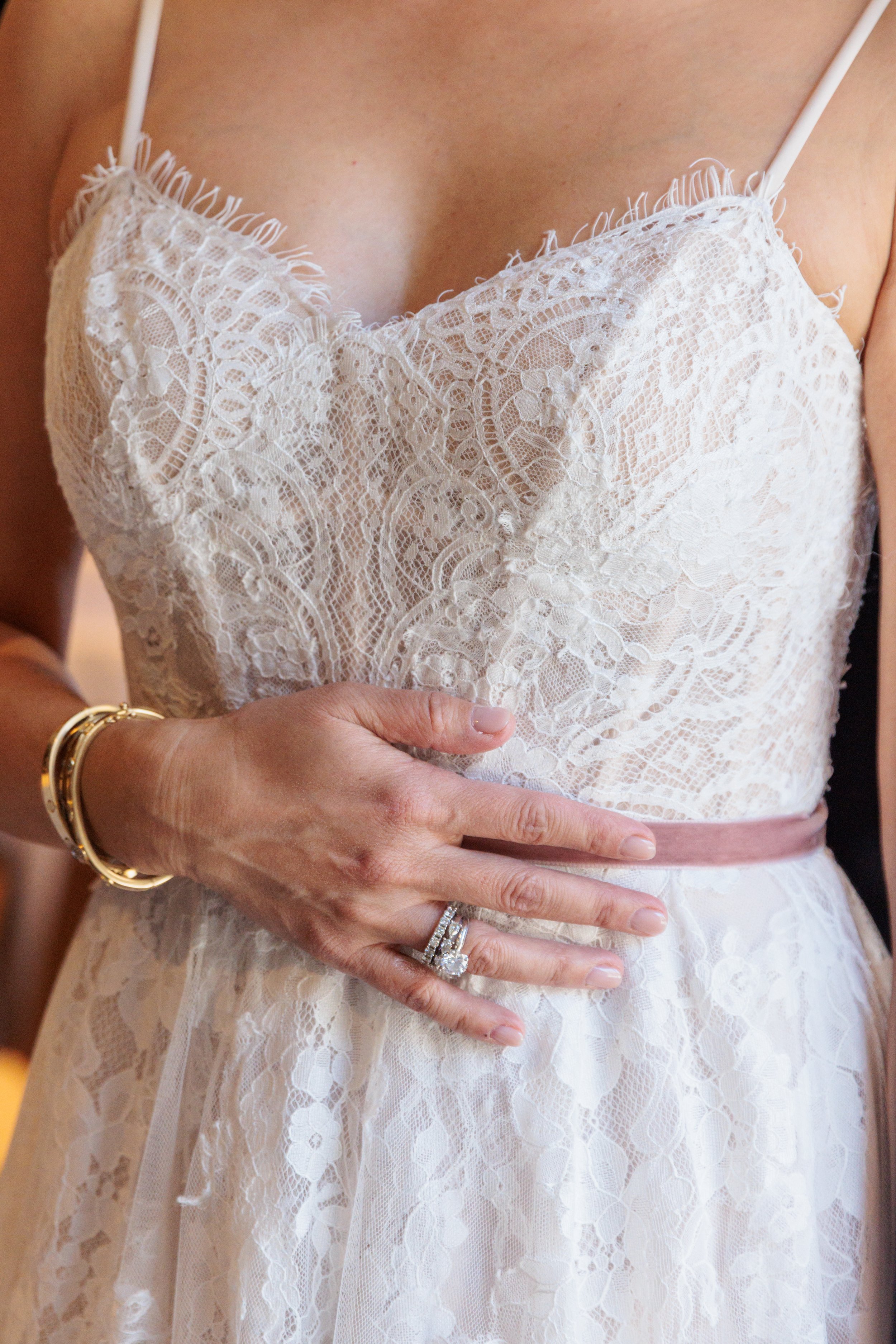 Close-up of a woman wearing a white lace dress, holding her stomach, with gold and diamond rings and bracelets visible on her fingers and wrist.