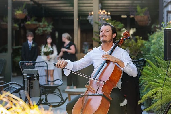 A man playing the cello at an outdoor event with a wedding party in the background.