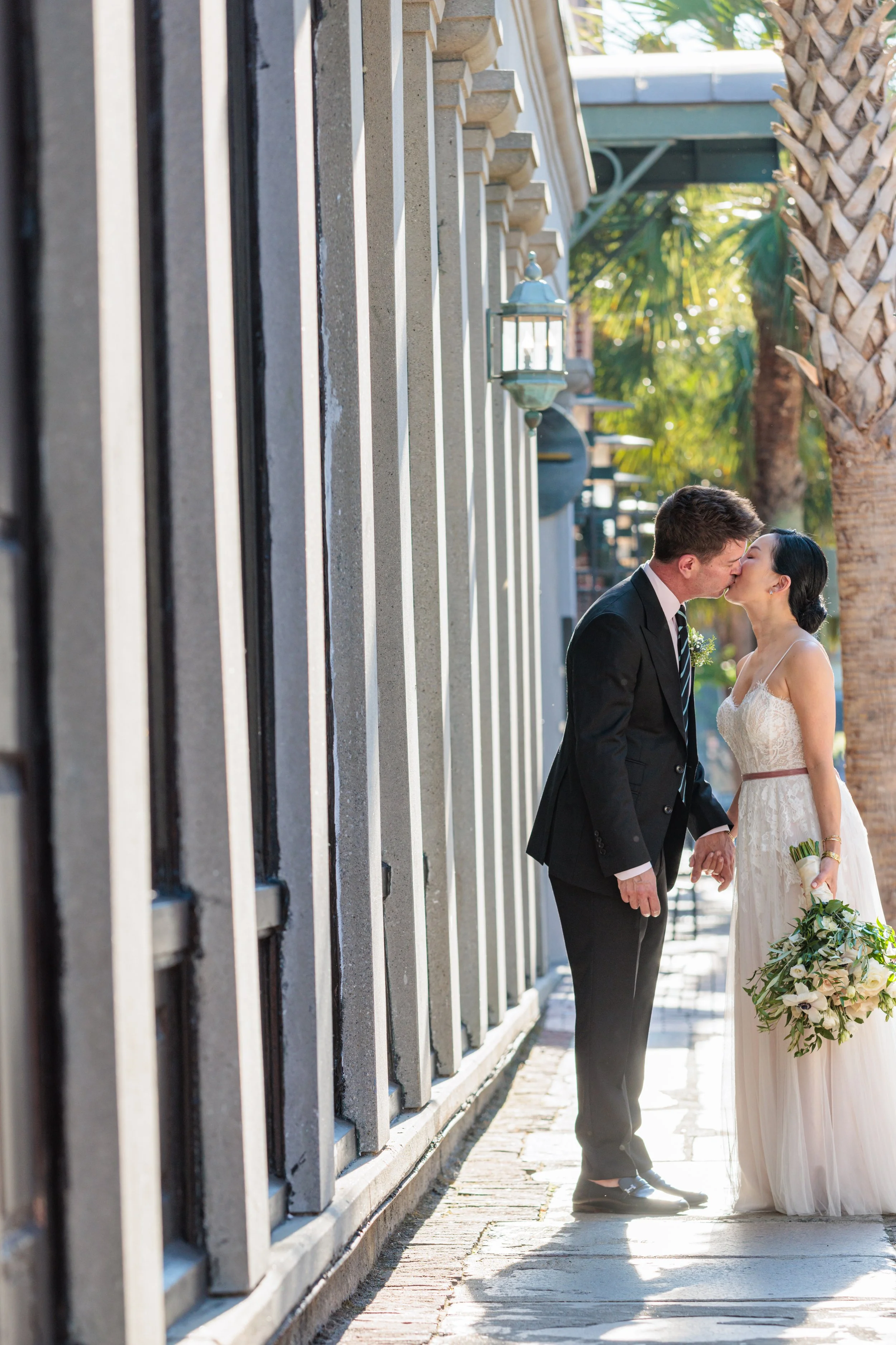 A couple in wedding attire sharing a kiss outdoors on a sunny day, with trees and decorative lampposts in the background.