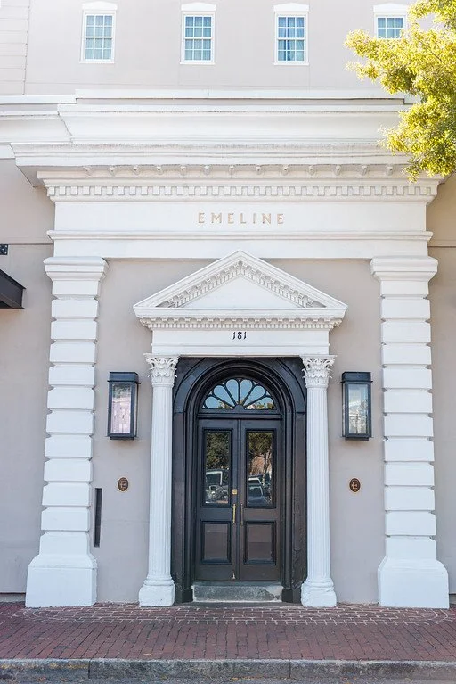 The entrance of a building with classical architectural elements, including white columns, a triangular pediment, and decorative moldings, with 'EMELINE' inscribed above the door and the number 181 displayed above the entrance.