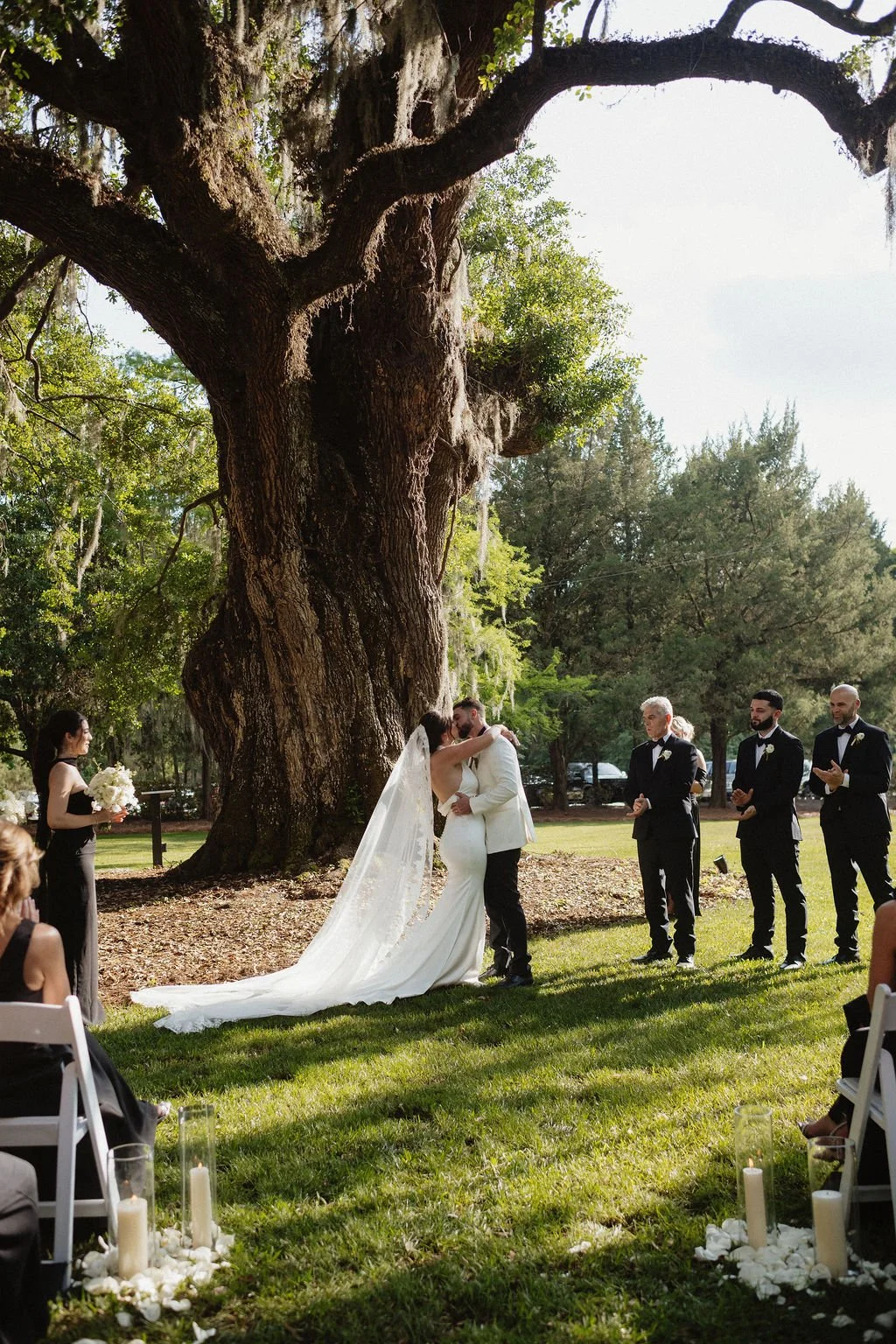 A couple in wedding attire share a kiss during outdoor wedding ceremony under a large old tree, with officiant and groomsmen in formal suits watching, and guests seated nearby, candles on the ground.