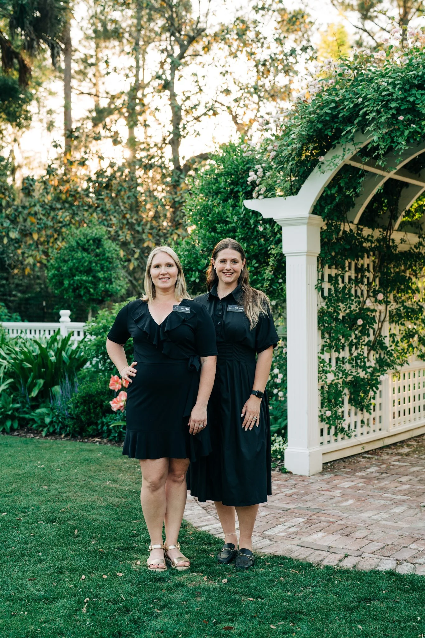 Two women standing outdoors in a garden, wearing black dresses and smiling. One has blonde hair and the other has brown hair. There is greenery and a white fence in the background.