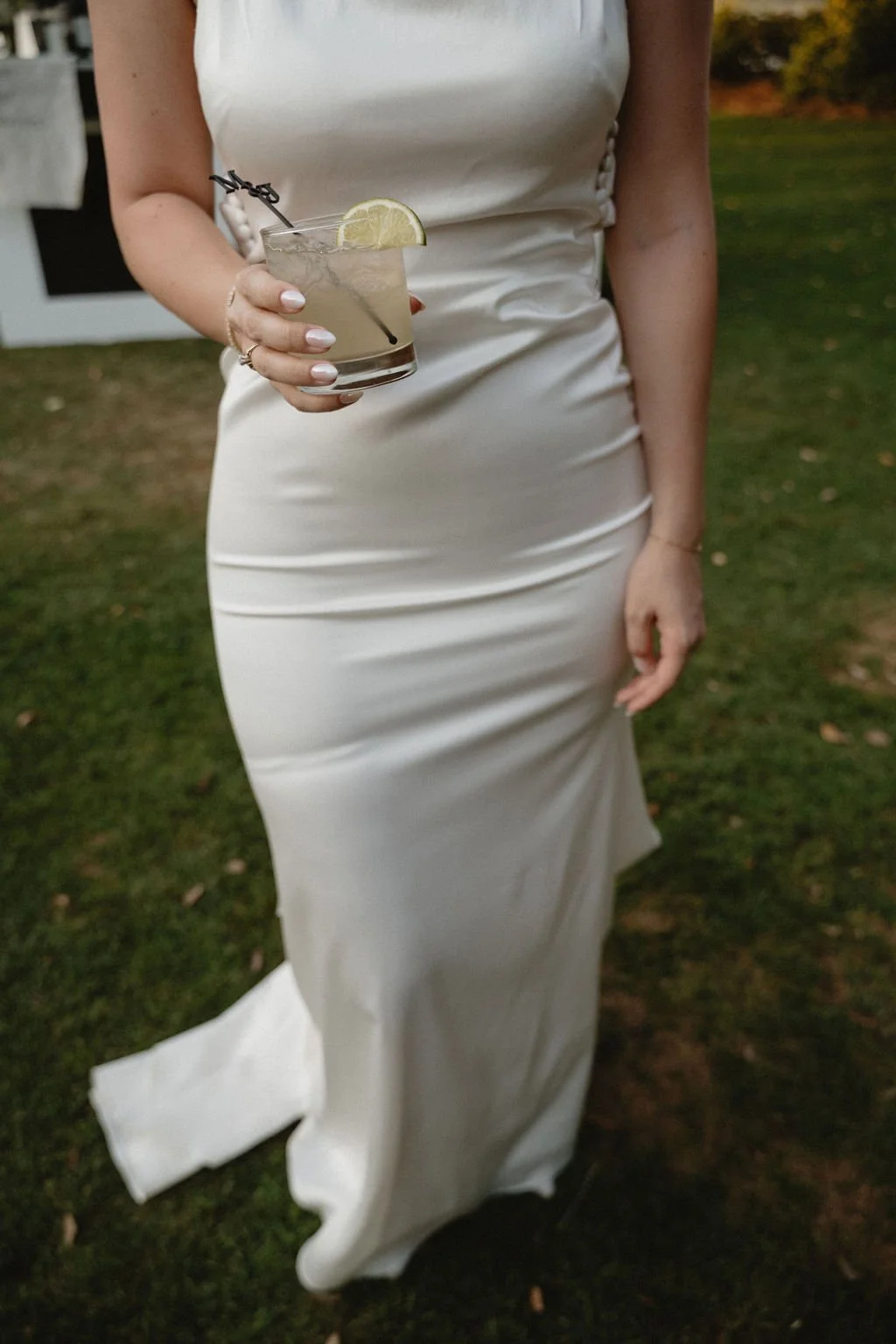 A woman in a white satin dress is holding a cocktail glass with a lemon wedge and a straw, standing outdoors on grass with a blurred background.