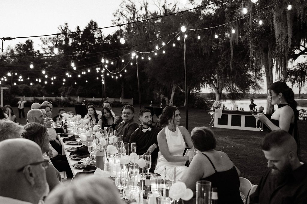 A dinner party outdoors during evening, with guests seated at a long table decorated with flowers, candles, and glasses, under string lights, near a body of water with trees in the background.