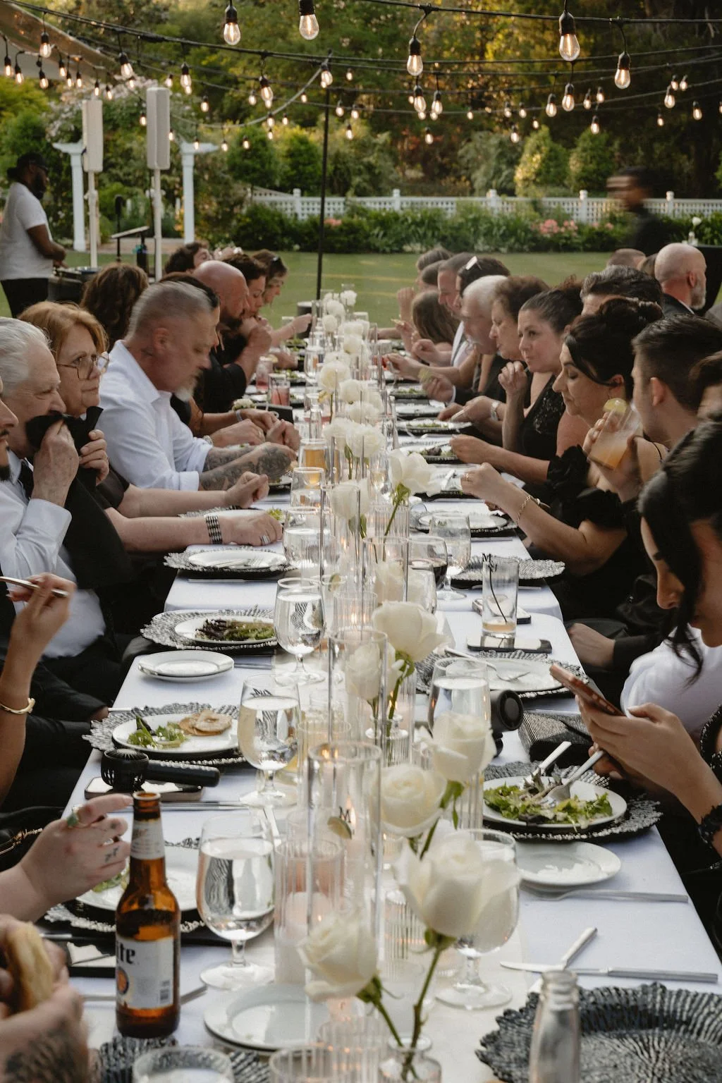 People gathered at a long outdoor dinner table decorated with white flowers and candles, enjoying a meal under string lights in a garden setting during evening.
