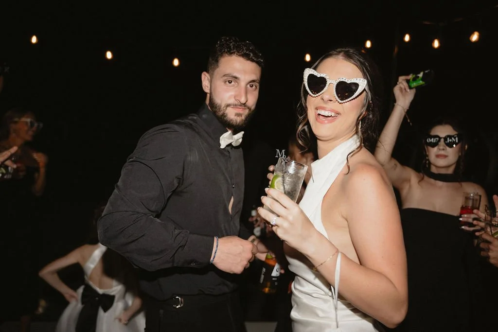 People dancing at a party with string lights, partygoers wearing sunglasses, and drinks in hand.