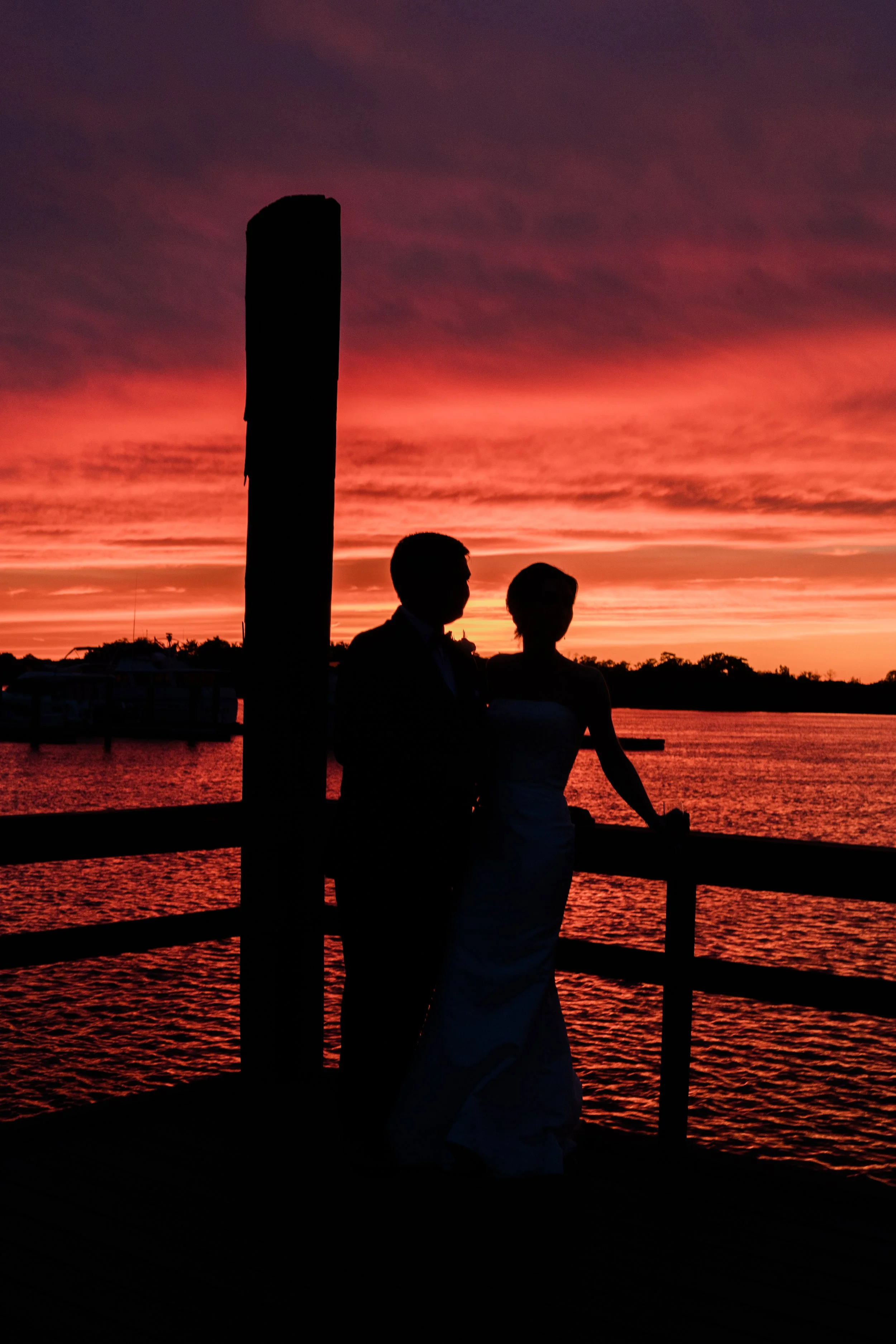 Silhouettes of a man and woman standing on a pier during a vibrant sunset with pink, purple, and orange clouds reflected in the water.