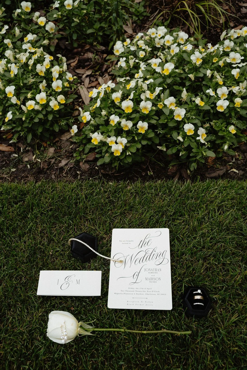 Wedding invitation, rings, necklace, and a white rose with a ring on it laid on grass at the base of a bush with white and yellow flowers.