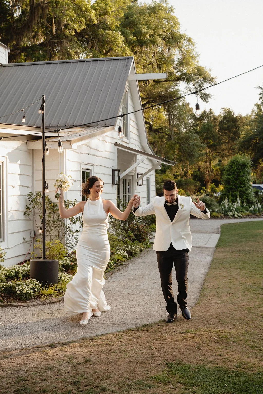 A bride and groom dancing outside their wedding venue, holding hands and smiling. The bride is wearing a white wedding gown and holding a bouquet, while the groom is dressed in a white tuxedo jacket and black pants. The setting is outdoors with a hou