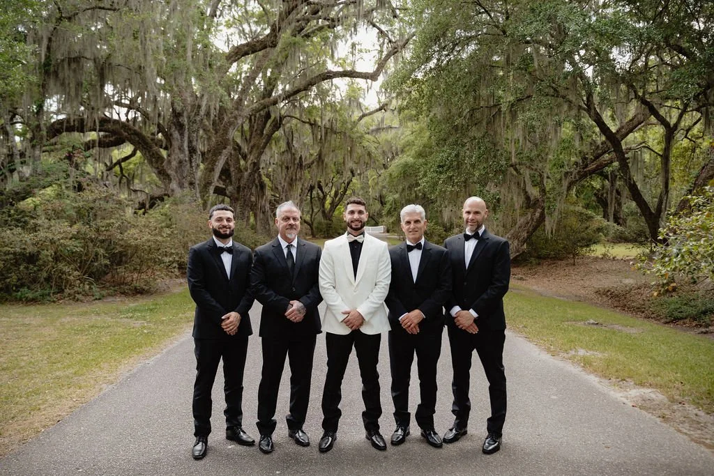 Groom and four groomsmen standing outdoors on a paved path surrounded by trees and greenery.