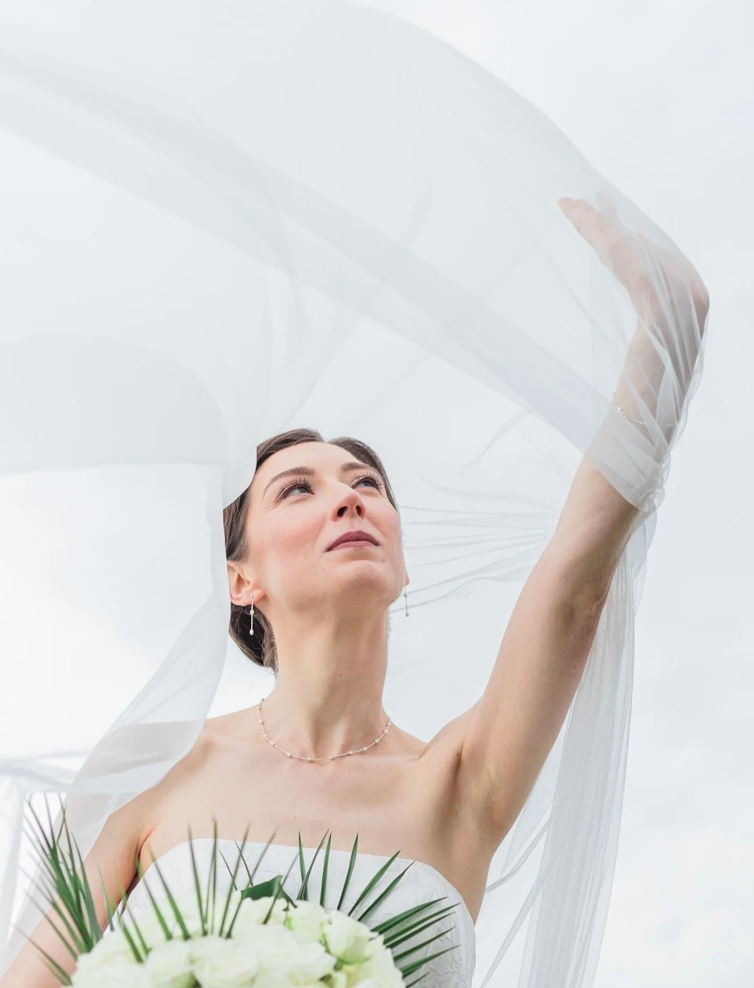A bride in a strapless wedding dress holding a bouquet of white flowers and green leaves, raising her arm and looking upward with a sheer veil flowing around her against a cloudy sky.