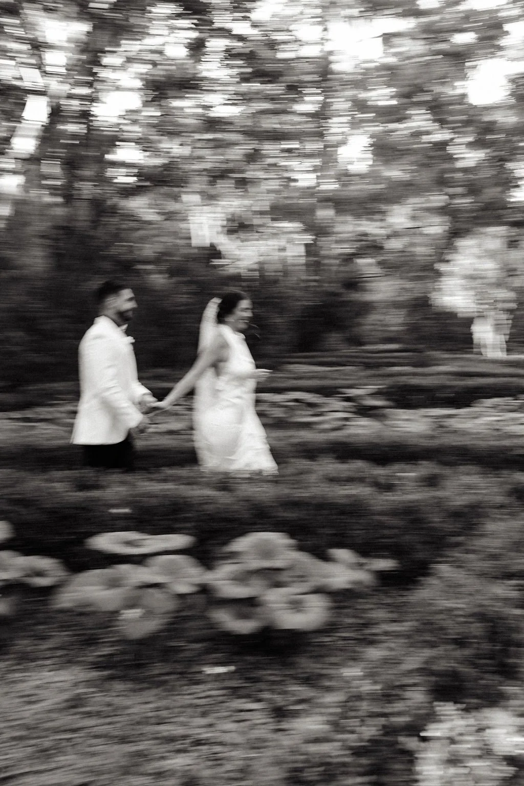 A blurry black and white photo of a bride and groom walking hand in hand outdoors with trees and flowers in the background.