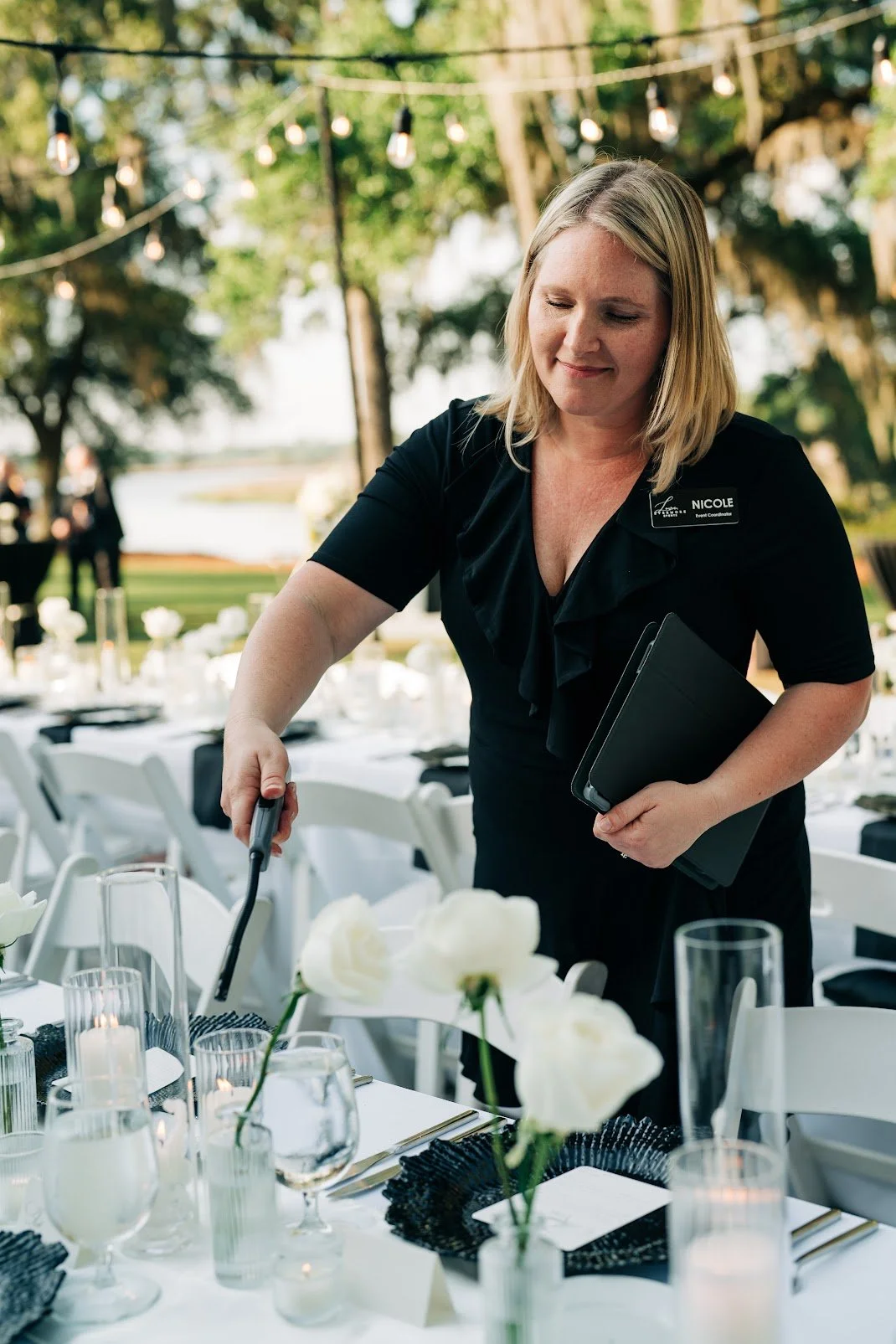 A woman with blonde hair, wearing a black outfit and a name tag, arranges a table with white flowers and elegant tableware outdoors, with string lights and trees in the background.
