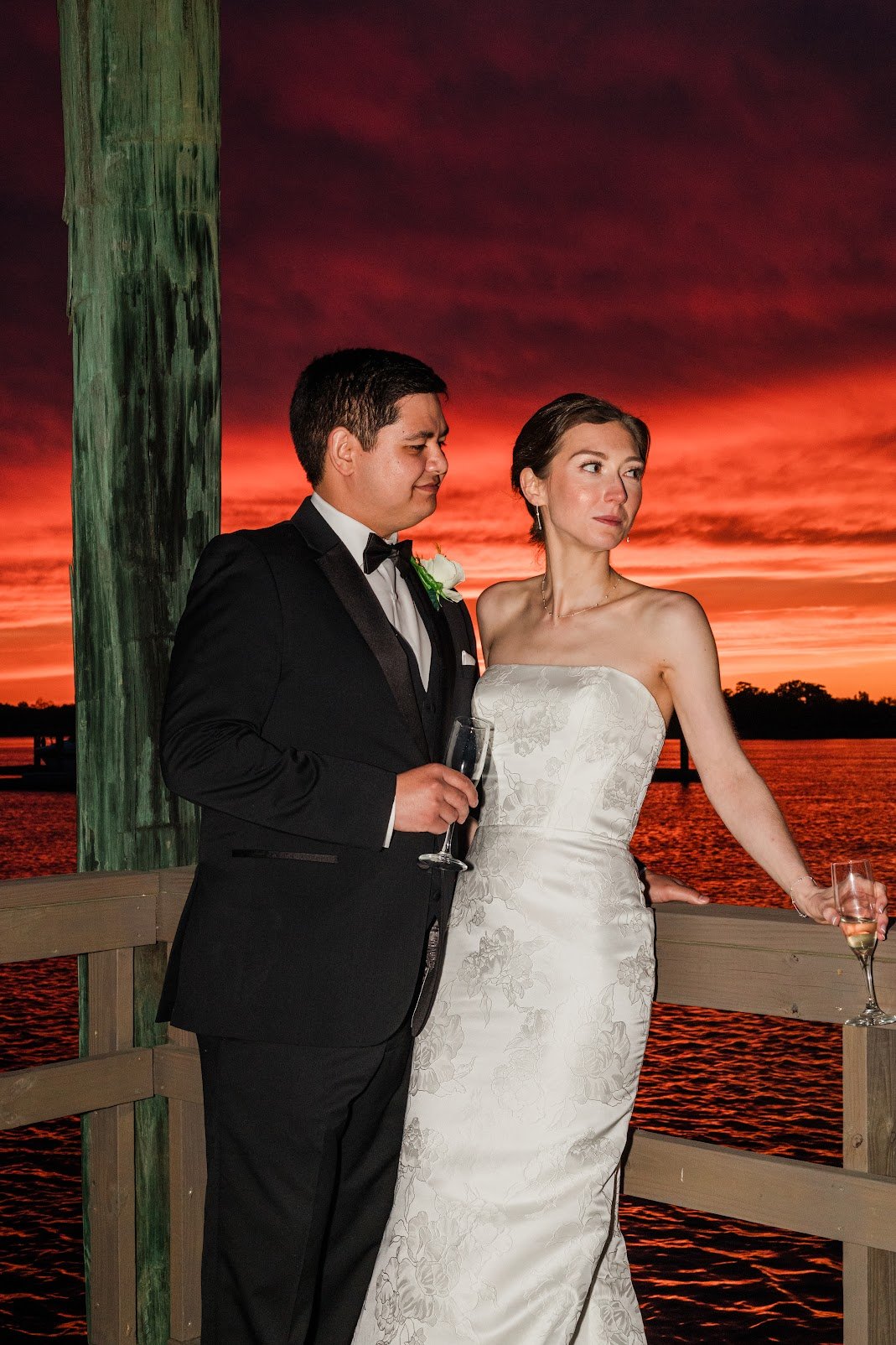 A newlywed couple in wedding attire standing on a wooden pier at sunset. The groom is wearing a black tuxedo holding a glass of champagne, and the bride is wearing a strapless white wedding gown, holding a glass of champagne, with water and a sunset in the background.