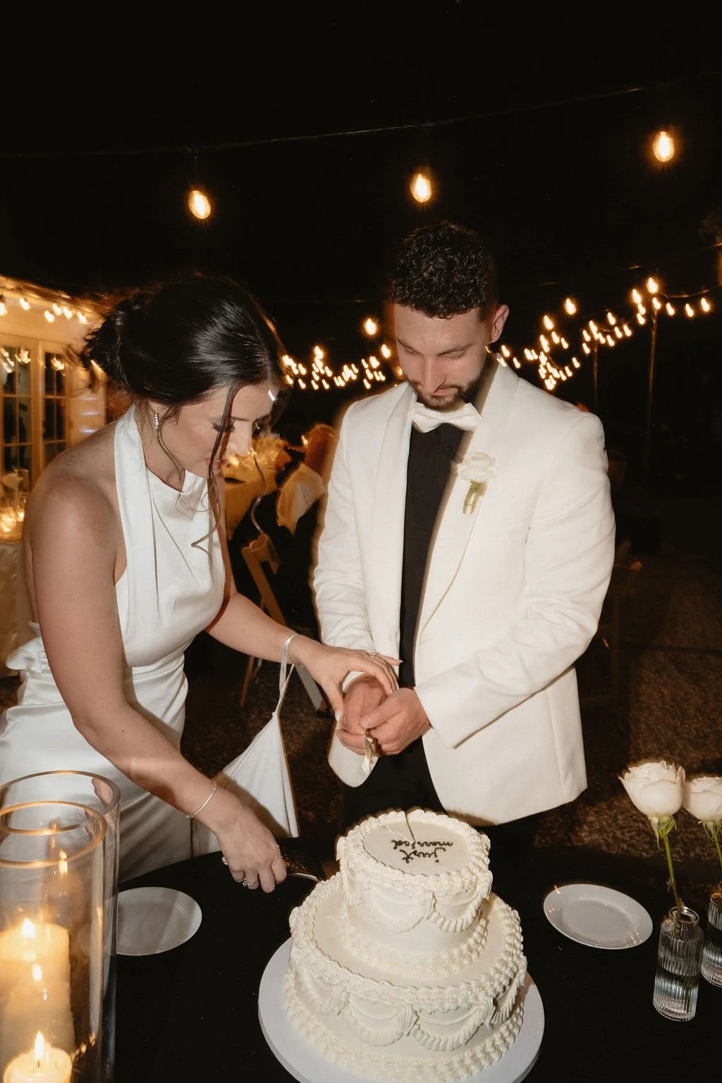 A bride and groom cutting their wedding cake at night under string lights, with candles on the table.
