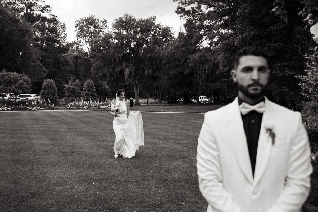 Black and white photo of a wedding couple outdoors; the man is in a white tuxedo with a bow tie, standing in the foreground and slightly blurry, while the woman in a wedding dress is in the background, walking on a lawn.