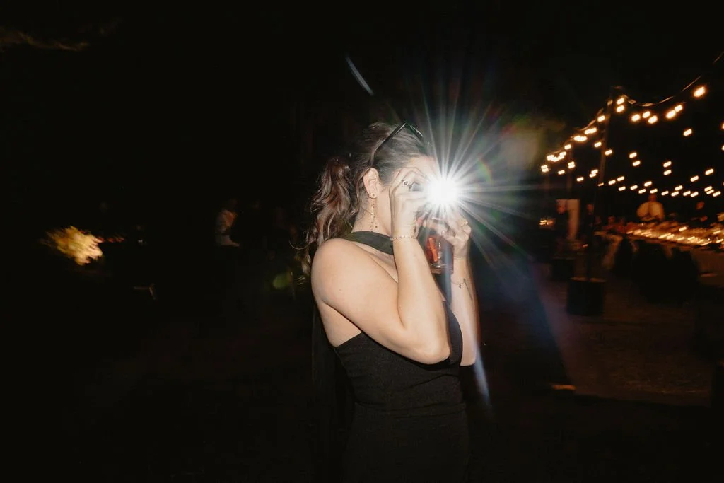 A woman in a black dress taking a photo with a camera at an outdoor event during nighttime, with string lights hanging in the background.