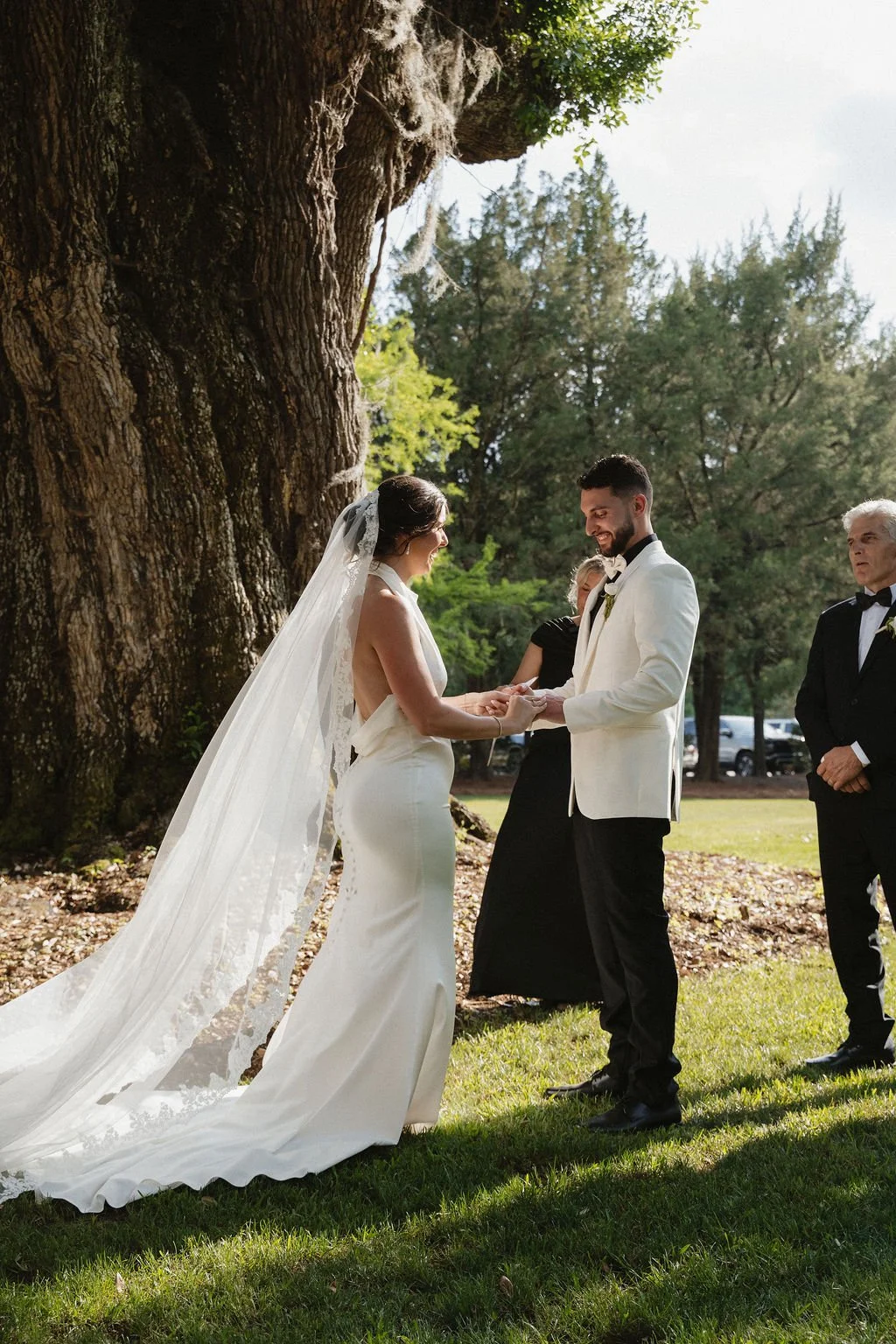 A wedding ceremony taking place outdoors under a large tree, with the bride and groom holding hands and smiling at each other, accompanied by officiants and guests.