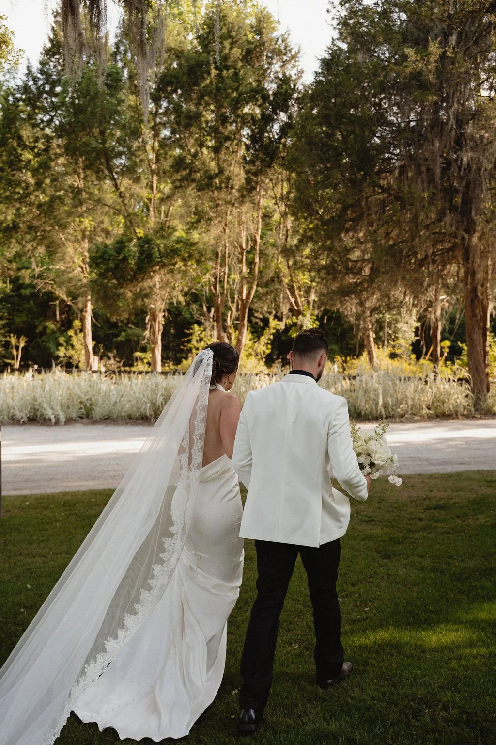 Bride and groom walking outside in a park during their wedding, with the bride wearing a long white gown and veil, holding the groom's hand, who is dressed in a white jacket and black pants, carrying a bouquet of flowers.