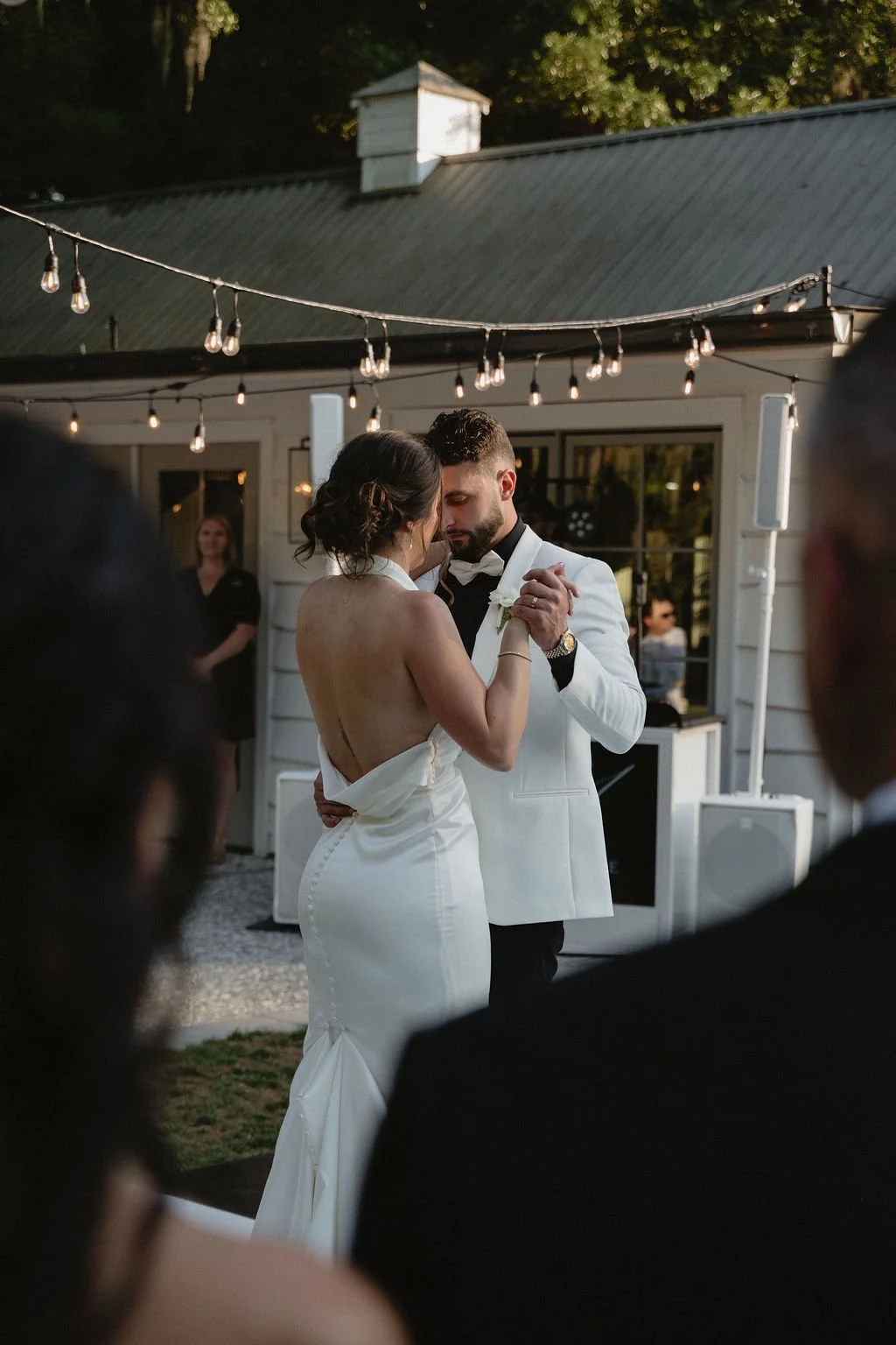 Couple sharing a dance at their wedding reception outdoors, surrounded by guests, hanging string lights, and a white building in the background.
